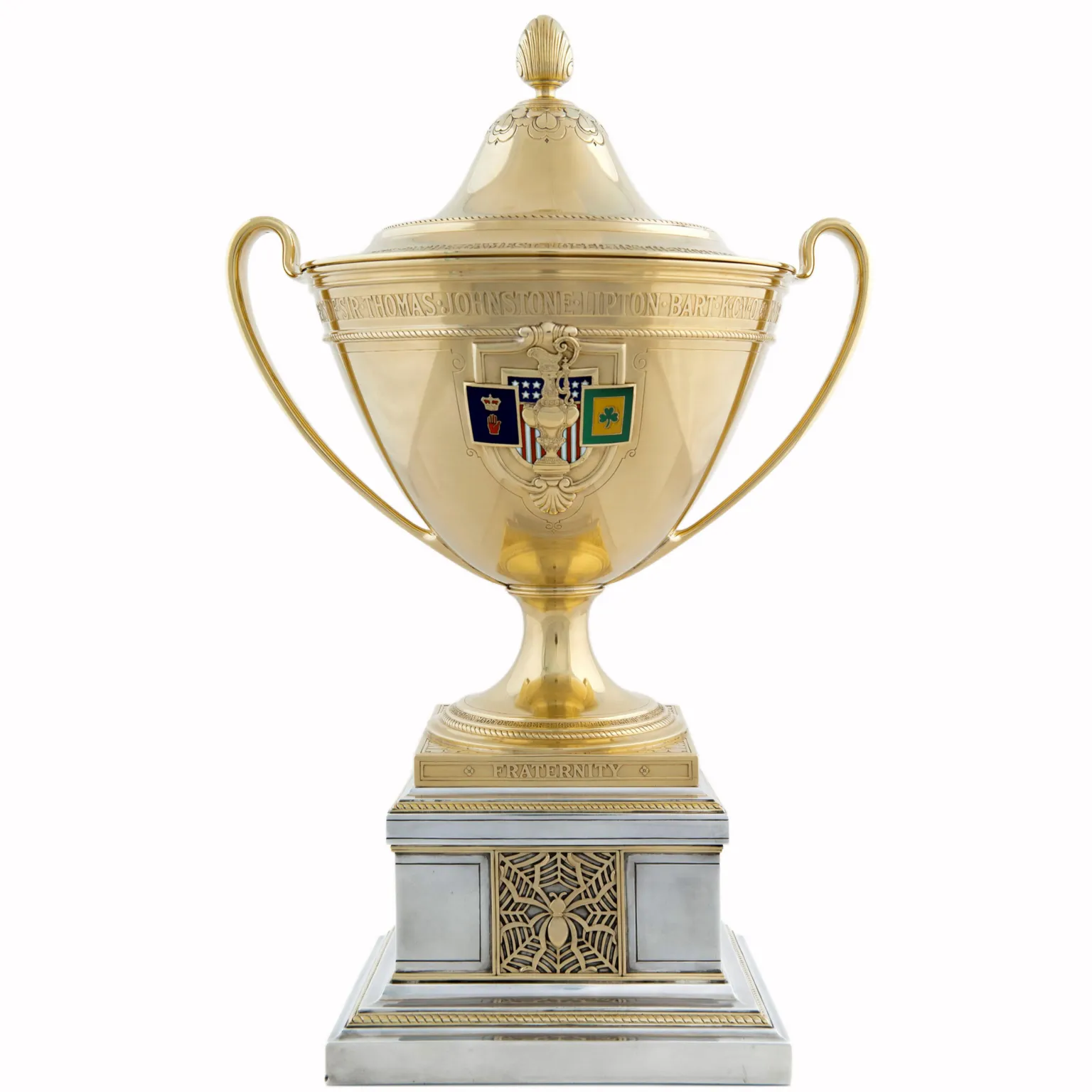 Glasgow Life Gold trophy cup with two curved handles and a lid, featuring an engraved inscription and a small shield emblem on the front, mounted on a decorative square base with the word “Fraternity” and a carved