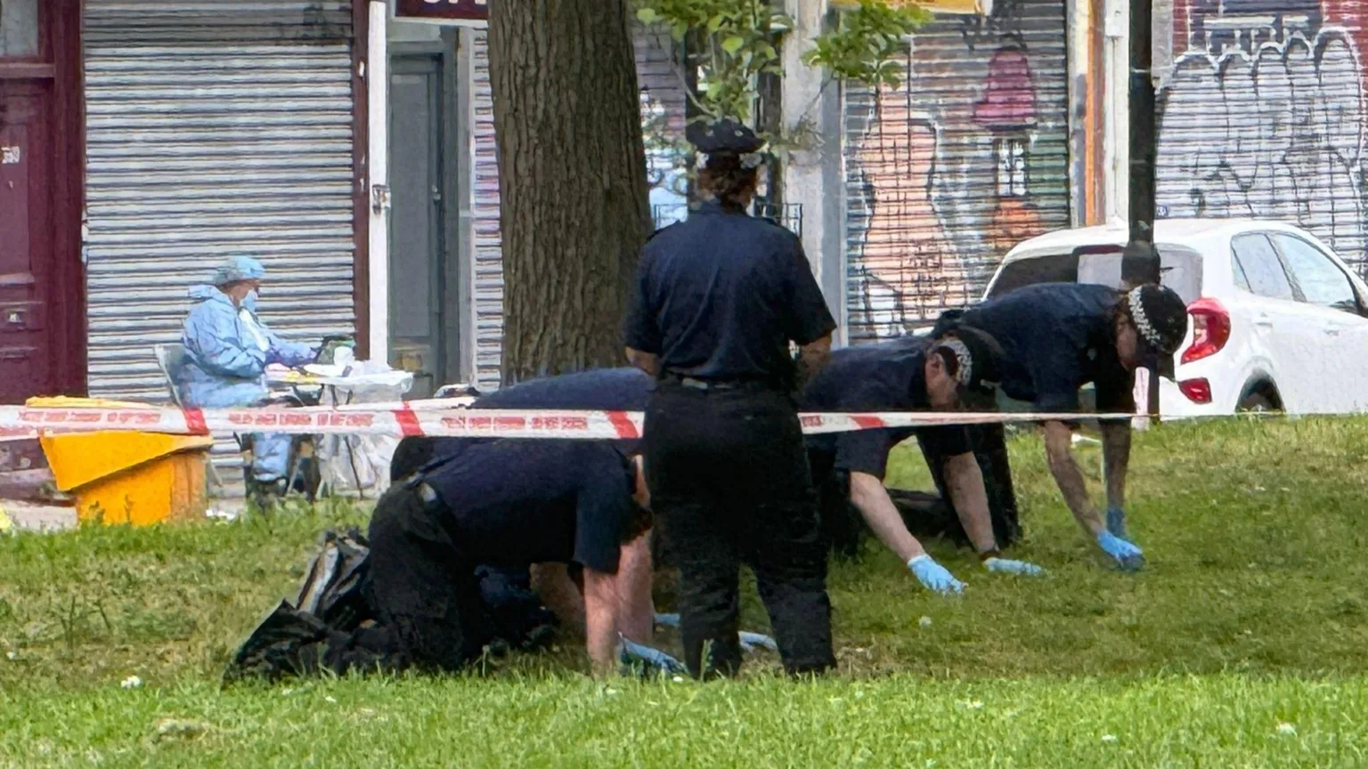 Rory Bibb/BBC Police officers hunched on the ground with blue plastic gloves going through the grass for evidence. You can see police tape in the front and in the back is a forensic officer. 