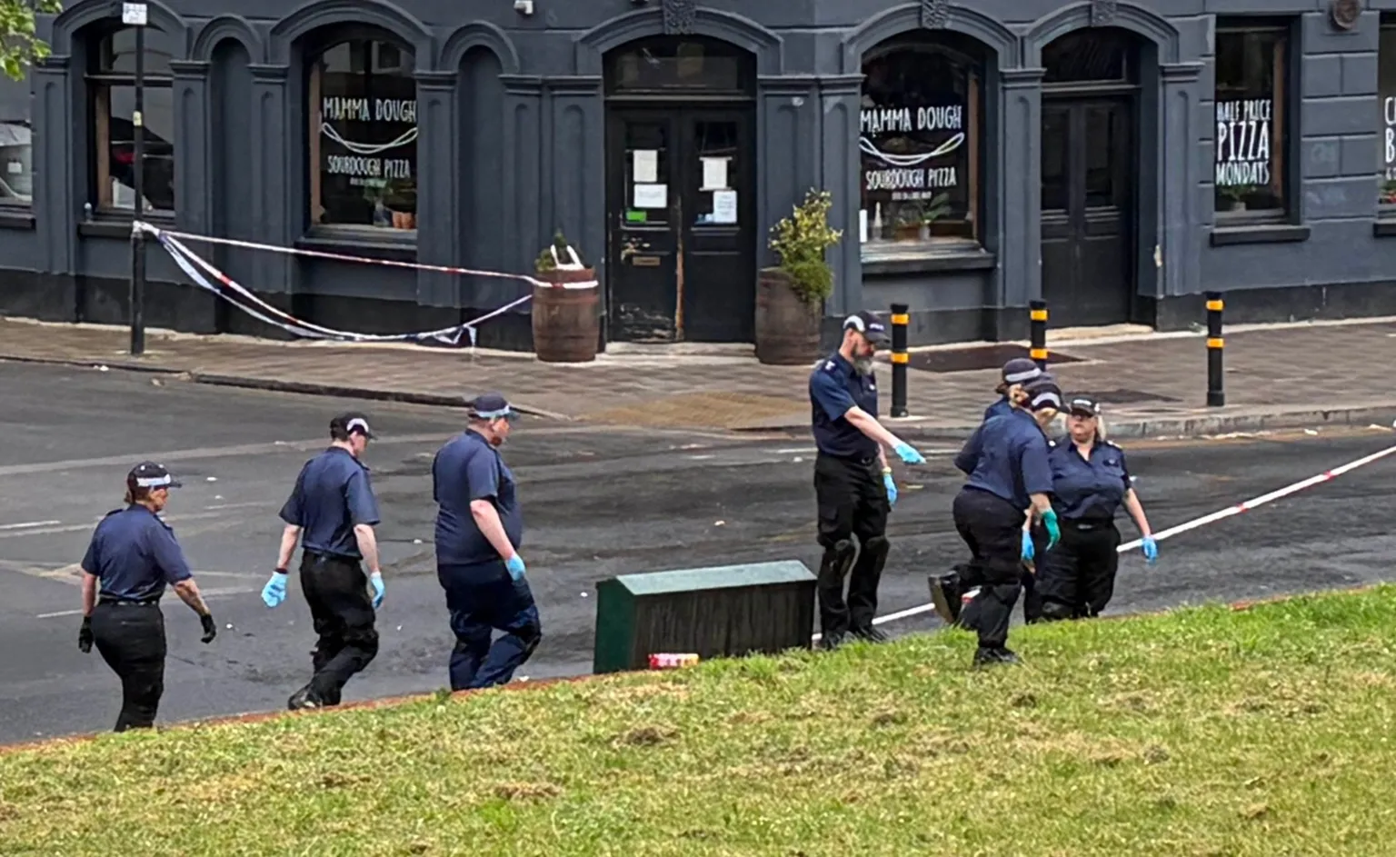 Seven police officers in short-sleeved blue shirts, black caps and blue surgical gloves search an area of grass. In the background, a grey restaurant frontage with 