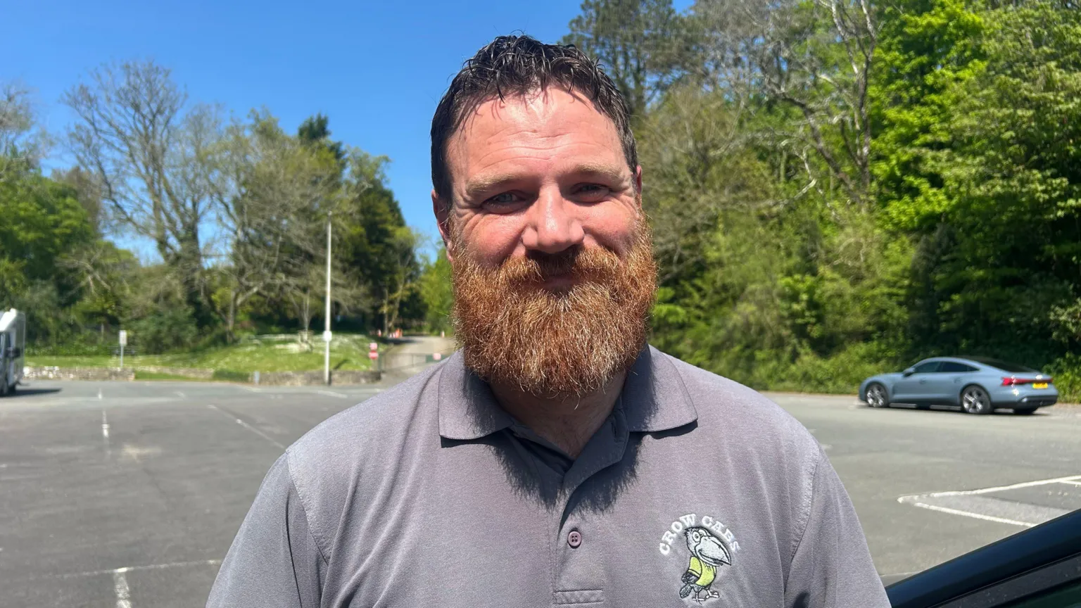  Steve Roberts in a car park. Wearing a grey polo neck with Crow Cabs logo on the right hand side. Has a light brown beard.