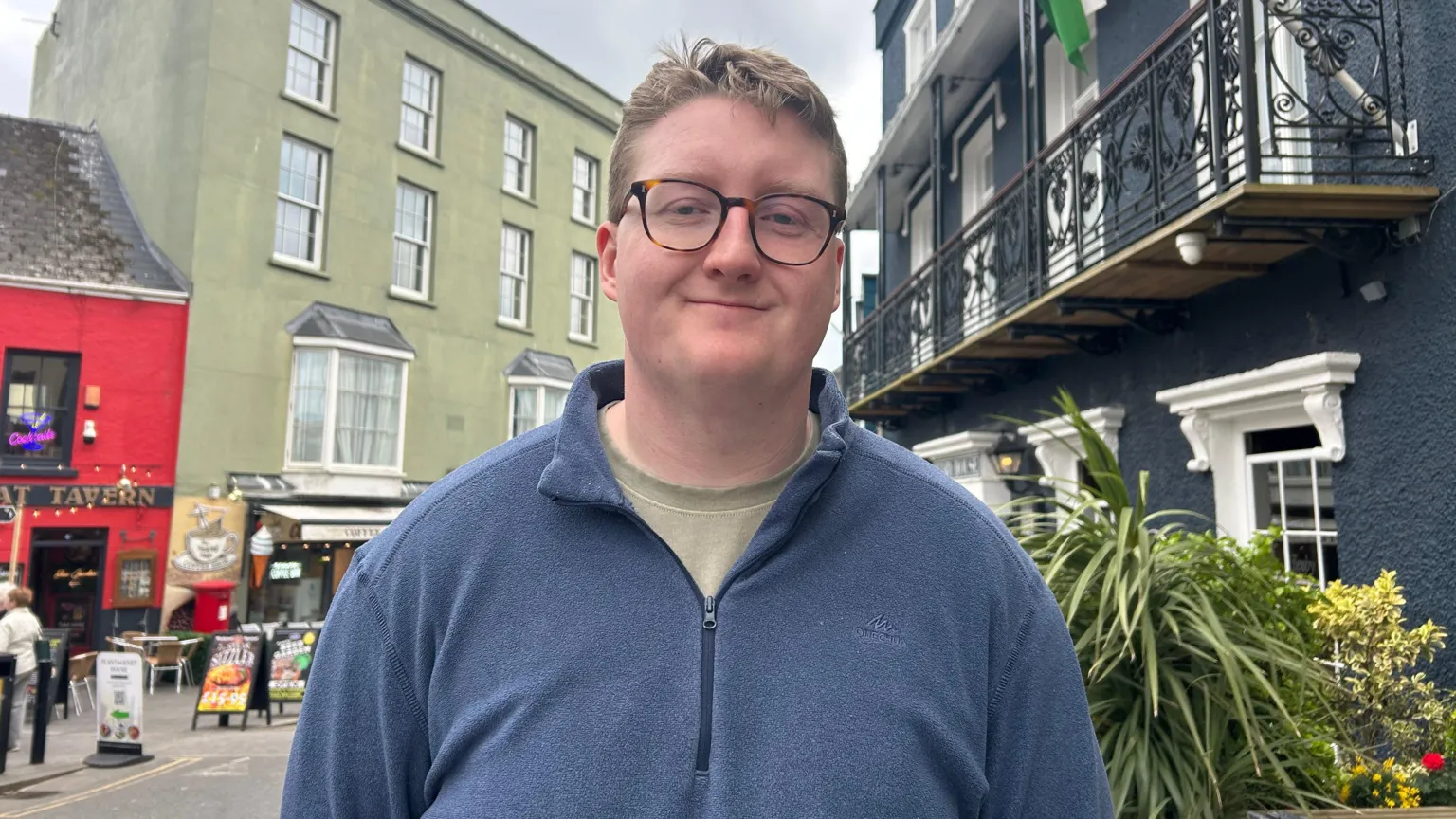  Frankie Bennet, wearing glasses and a blue quarter zip jumper and a light green t-shirt.