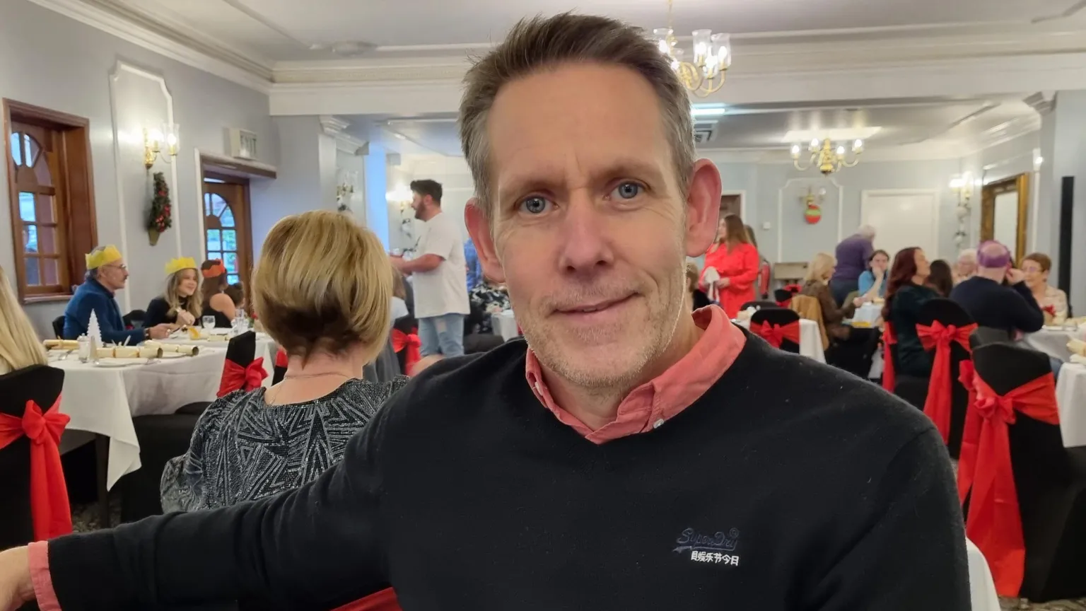 Tina Thomas A man with grey hair and blue eyes smiles straight into the camera. He is wearing a red shirt with a navy jumper on top, and is sitting in a function room, with other people sat at tables behind him