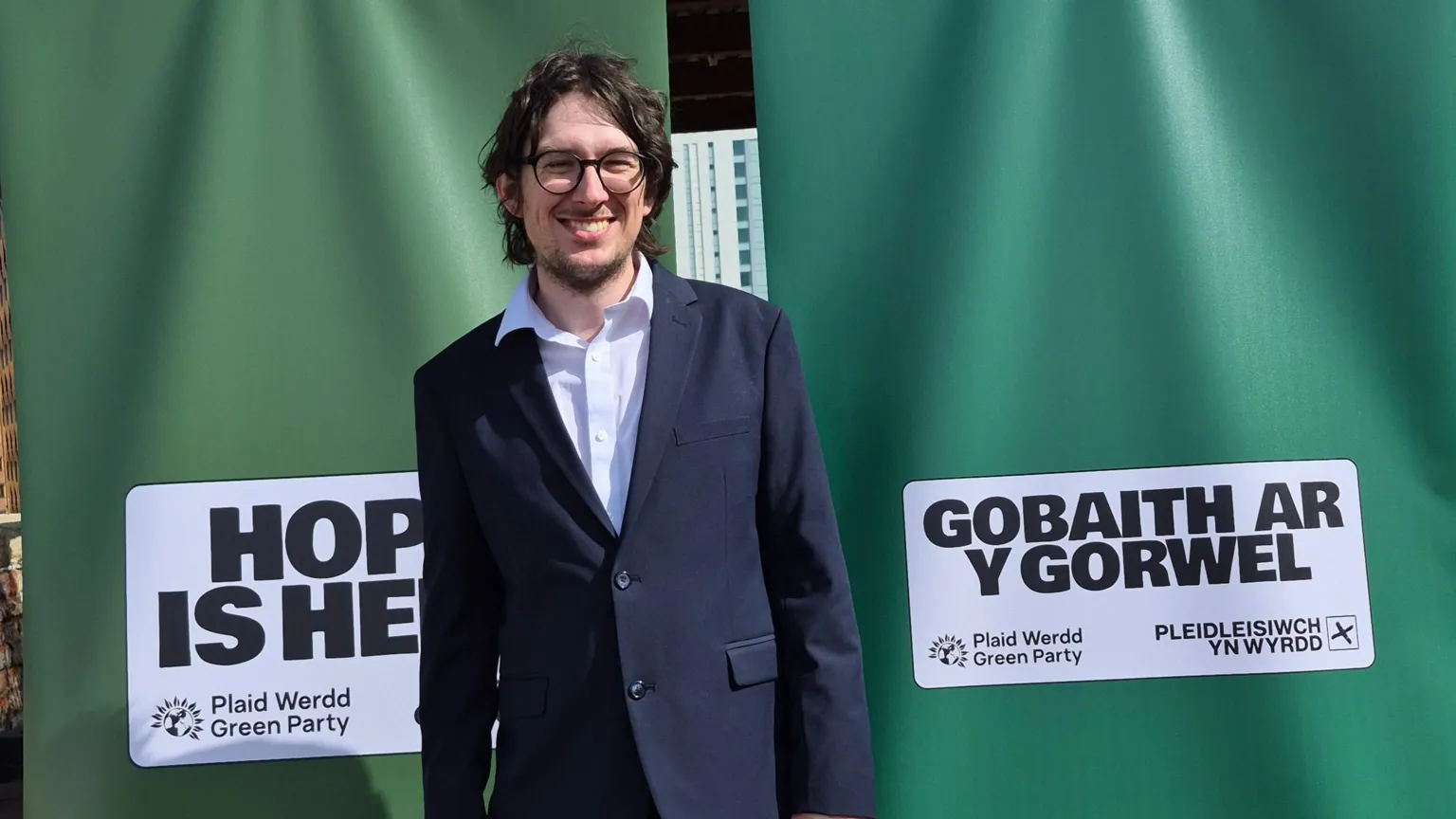 Angela Karadog A man in a navy suit and a white shirt, with dark hair and glasses stands smiling to camera. Behind him are two green banners with the Green party logo on. 