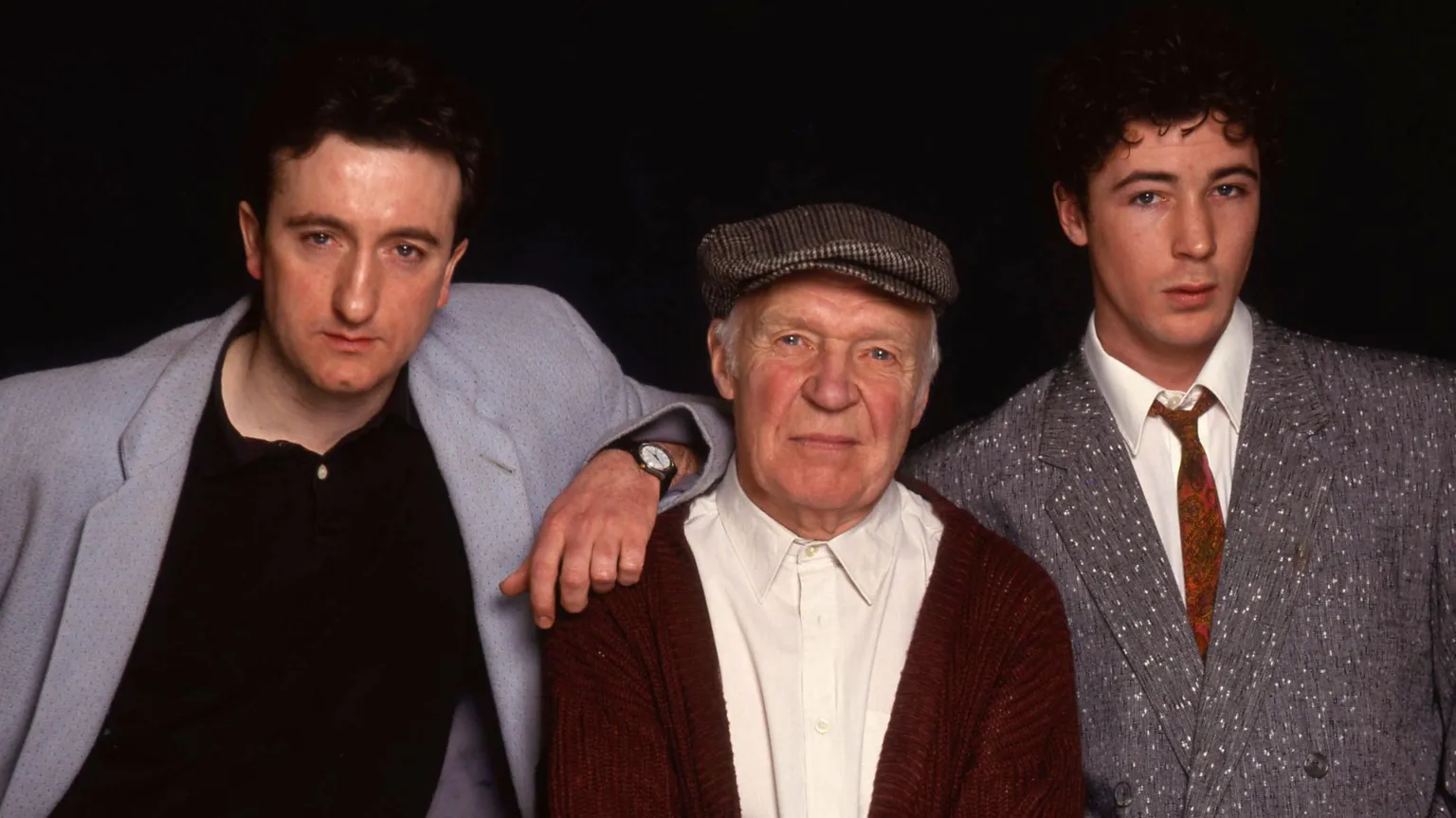Three men pose together against a dark background. An older man in a flat cap and red cardigan stands in the center with his arms folded, while two younger men stand on either side—one leaning in with his arm on the older man’s shoulder, and the other in a gray suit. All three look directly at the camera.