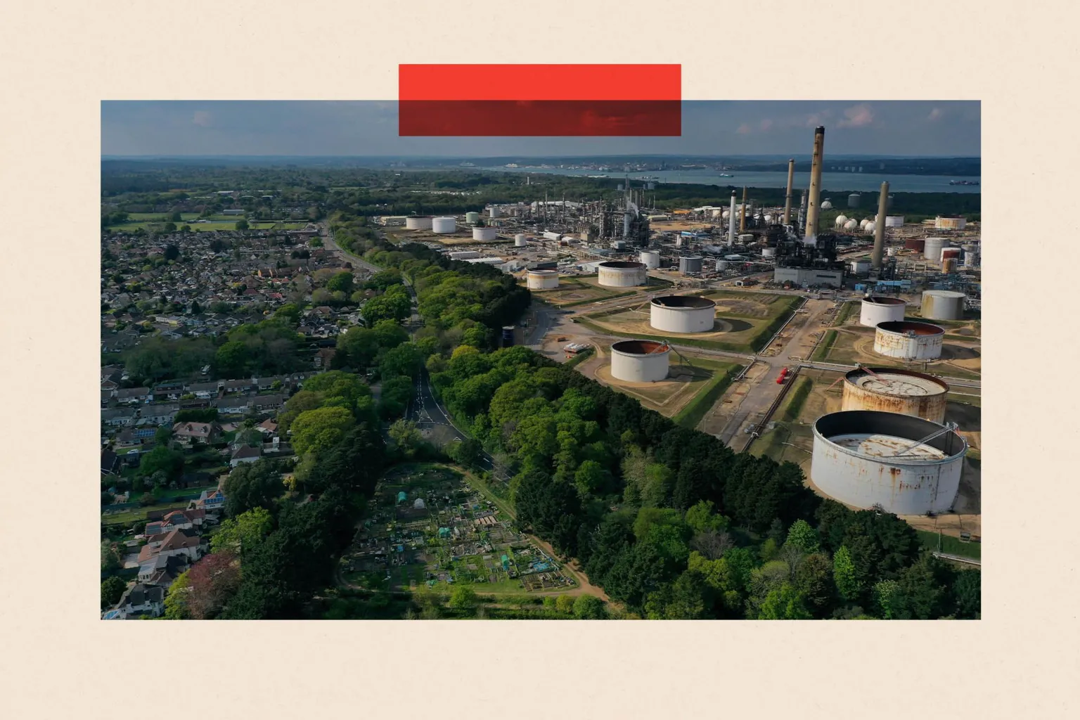  Aerial view of a large oil refinery with chimneys and circular storage tanks, bordered by woodland and a nearby residential neighbourhood, with water visible in the distance; a red rectangular graphic overlays the top of the image.