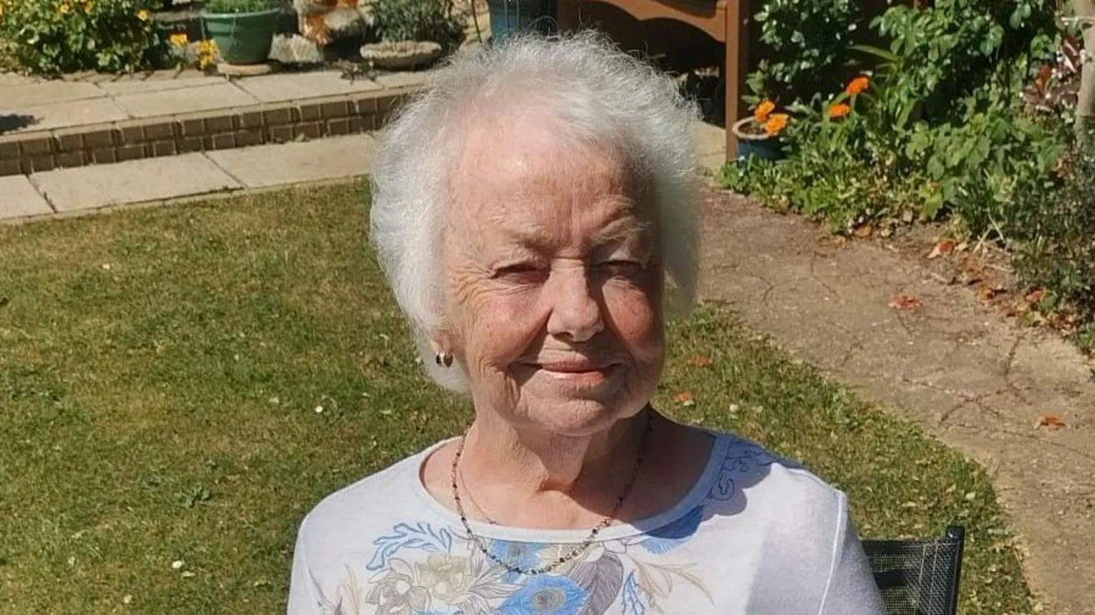 Colin Xerxes An older woman with white hair and a white and blue floral top is looking at the camera and smiling. She is sat in her garden with flowers and paving in the back of the picture. 