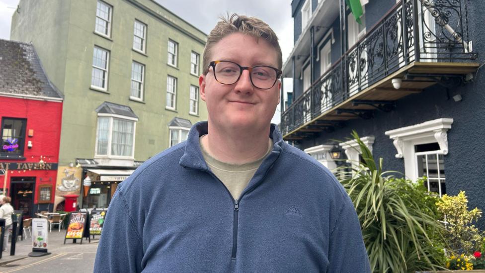 Frankie Bennet yn gwisgo crys-t gwyrdd a siwmper glas a sbectol