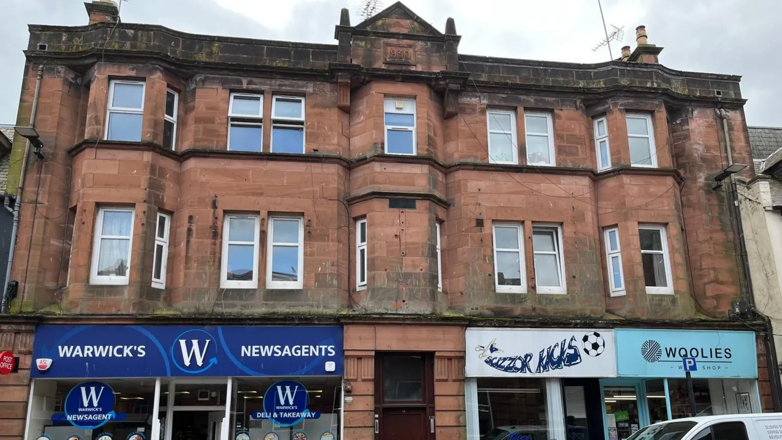  A large sandstone building with multiple windows and a shop on either side of a burgundy door on the ground floor 
