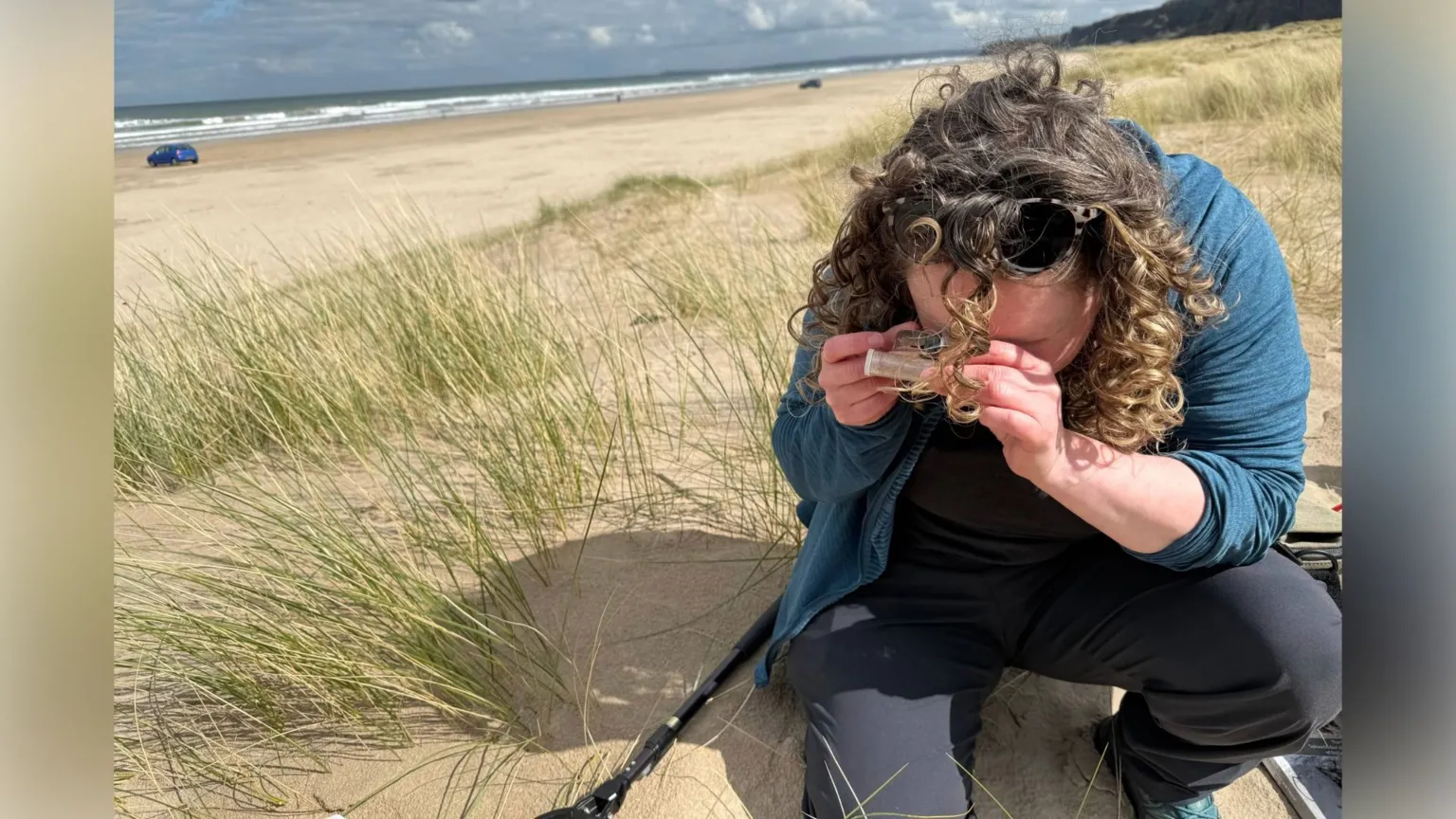 Mel wearing a blue jacket and black trousers looking closely at a beetle through a magnifying glass. A beach is behind her. She is sitting in the sand dune. 