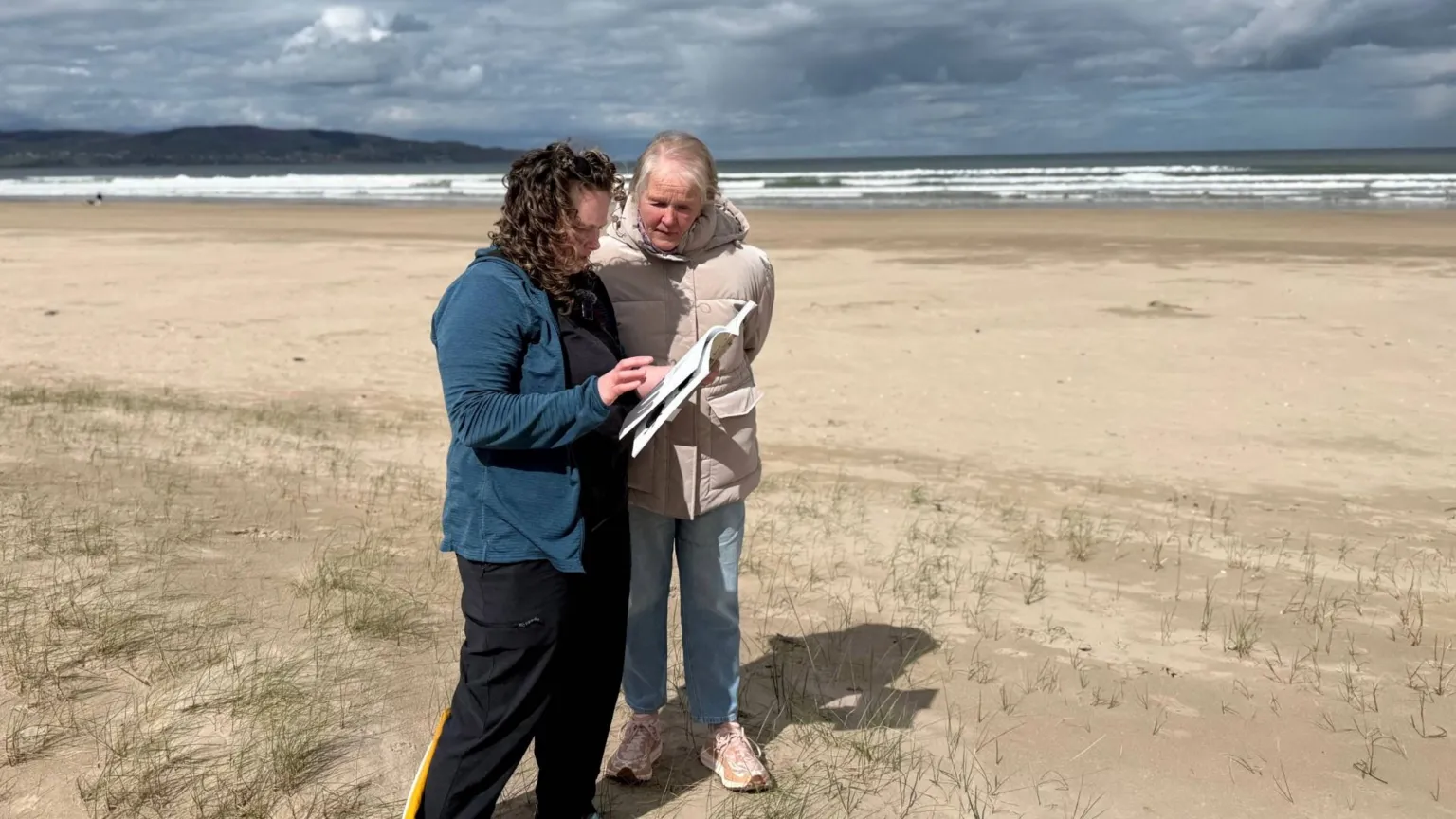 Mel speaking to a beech visitor about beetle surveying. Mel is wearing a blue jacket and black trousers. The visitor is looking at a book on beetles that Mel is holding. The visitor is wearing a white coast. They are both standing at the edge of the sand dunes on Benone beach. The sea is also in the background. 