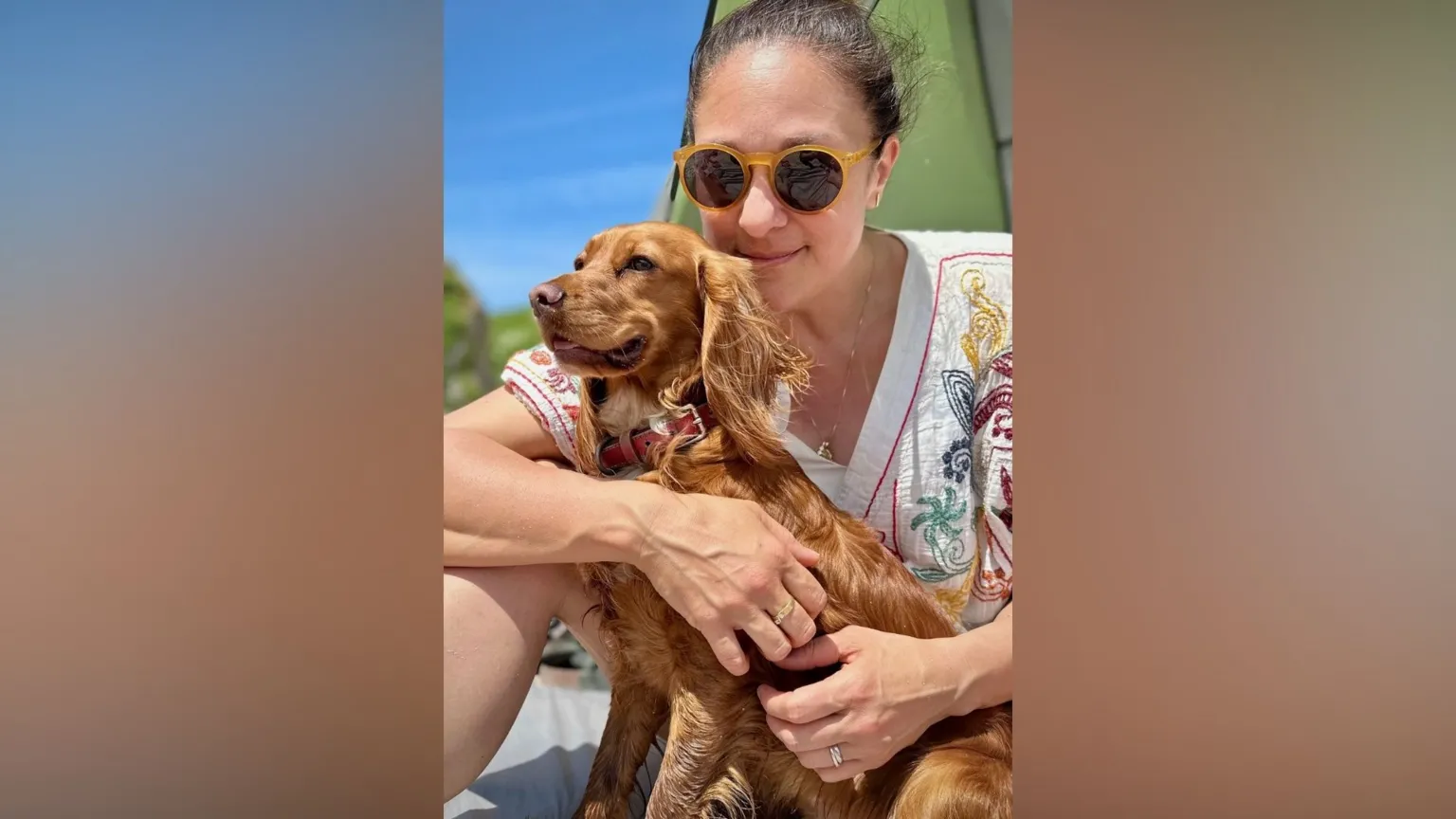Shelley Perkins Shelley Perkins smiles at the camera as she holds her dog Roxy, a golden cocker spaniel. Shelley has dark brown hair and is wearing sunglasses and a floral top.