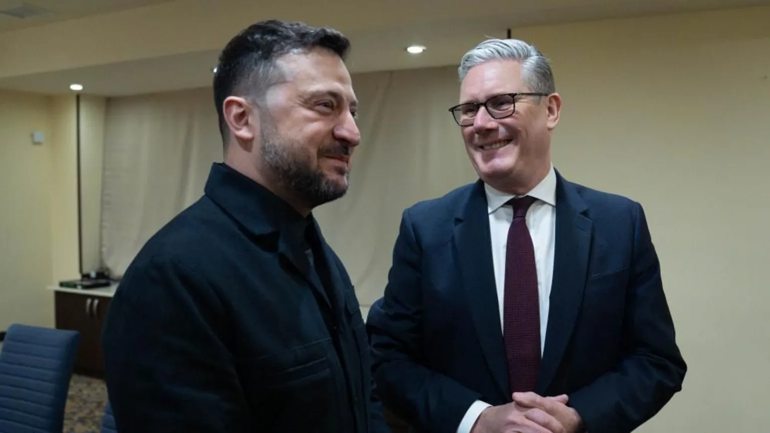  Sir Keir Starmer is stood on the right and is smiling as he looks at Ukrainian President Volodymyr Zelensky. The two men are stood in a hotel conference room which has white walls. Sir Keir is wearing a dark coloured suit jacket, a white shirt, a dark red tie and glasses. Zelensky is dressed in black.