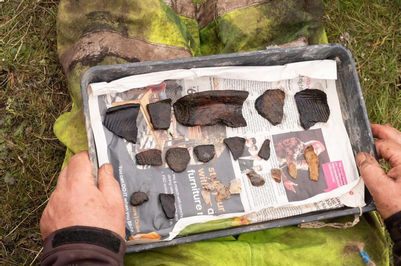  A pair of hands holds a tray containing broken pieces of ancient pottery