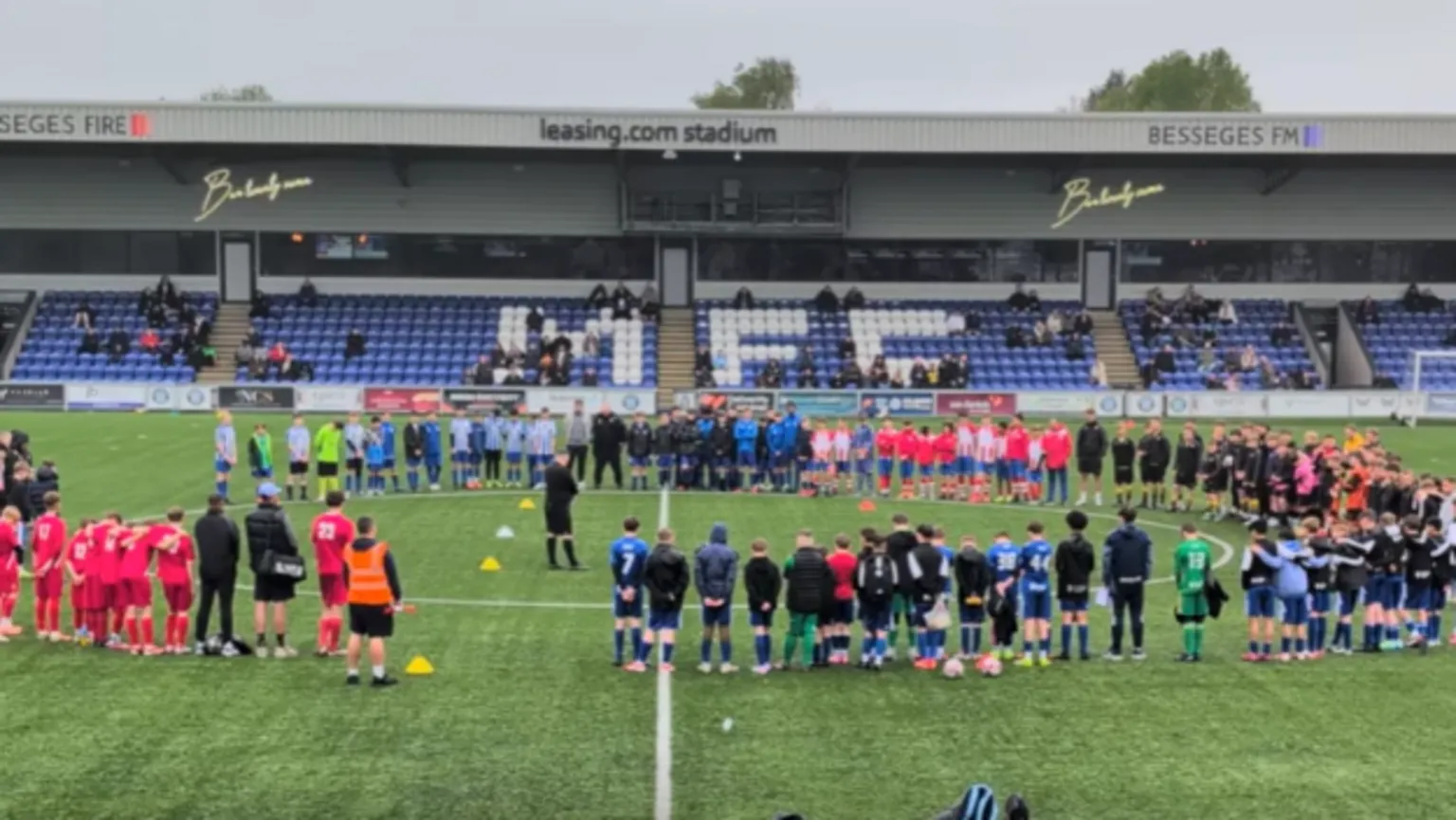 Football 4 Macclesfield Young footballers and adults connected to the team and summer camp stand in a circle on a pitch paying tribute to Brodan. They are wearing many different coloured kits and a referee appears to be standing in the middle of the circle. 