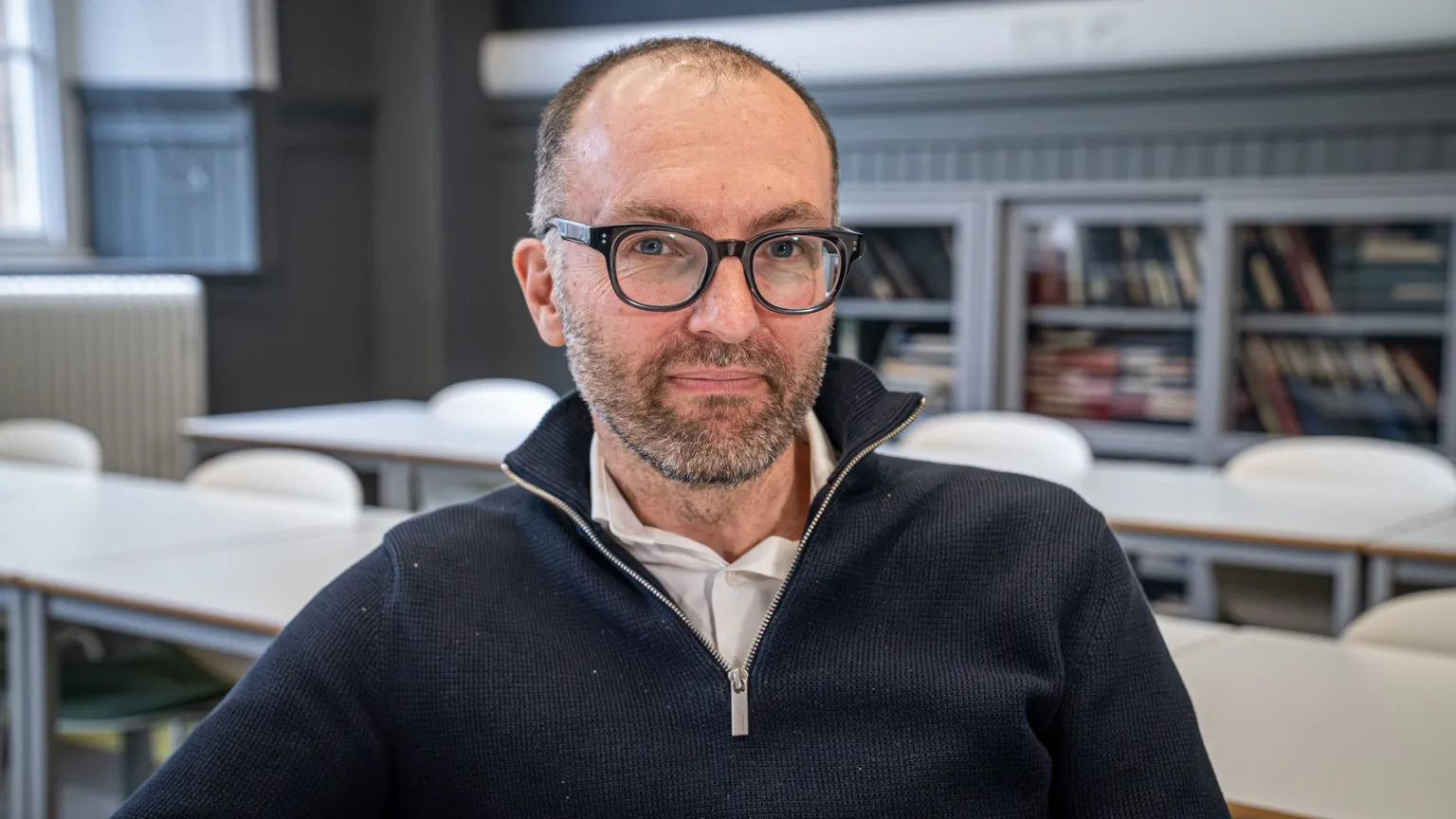 Prof Jamie Pearce, photographed in a lecture room at the University of Edinburgh. Behind him are rows of white desks, a large grey bookcase stretching the length of the room and windows to the left. He is wearing a navy blue three-quarter zip jumper, with a white shirt underneath. He is also wearing dark-rimmed glasses and has short, brown hair and a short, grey and brown beard. He is slightly smiling at the camera.