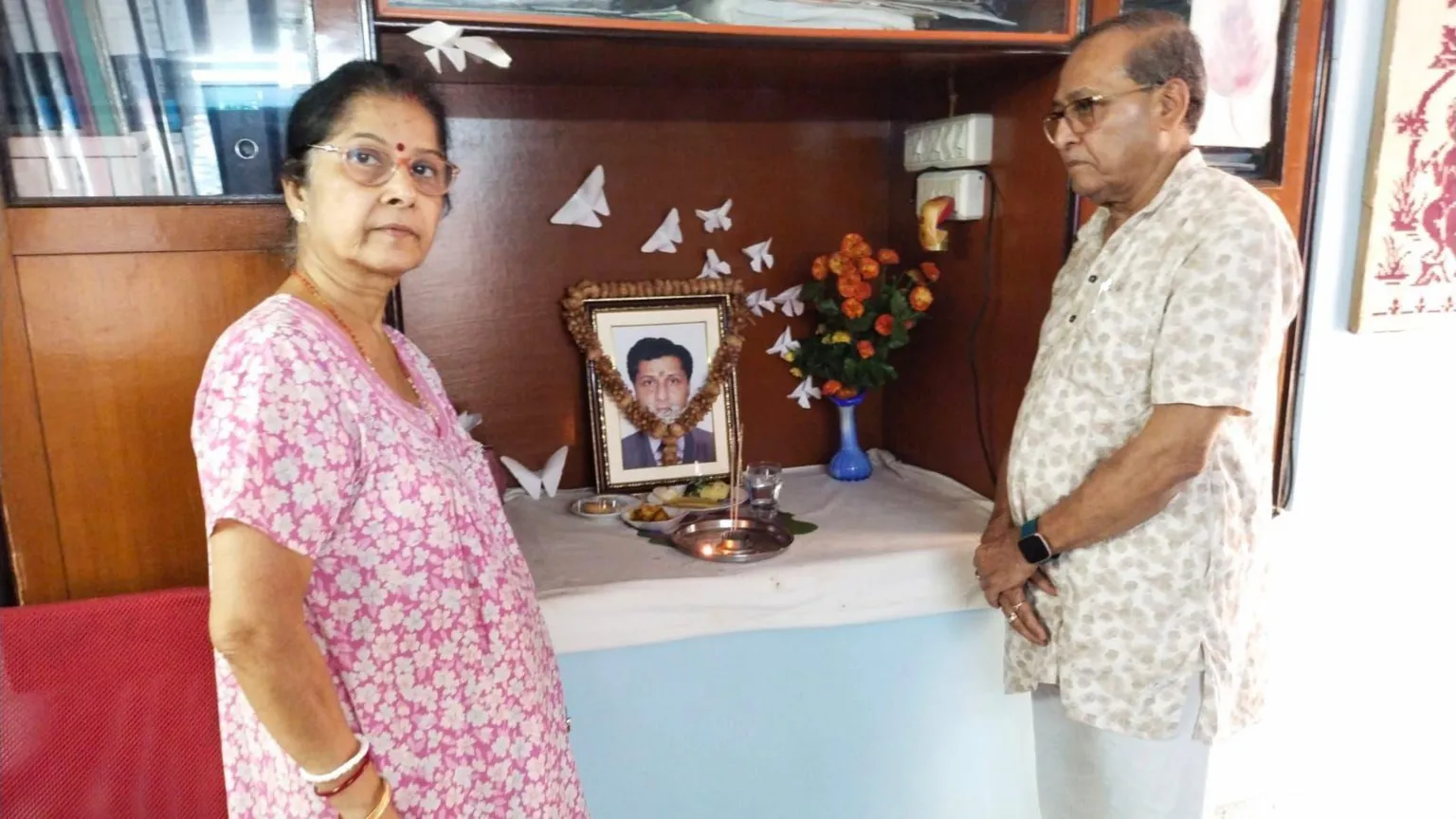 Sapna Mukherjee Sougat's mother Soma, 70, and his father Syamal, 79, standing beside a small altar in their home. At the centre is a photograph of Sougat. There are also orange flowers in a vase and what looks like white origami birds