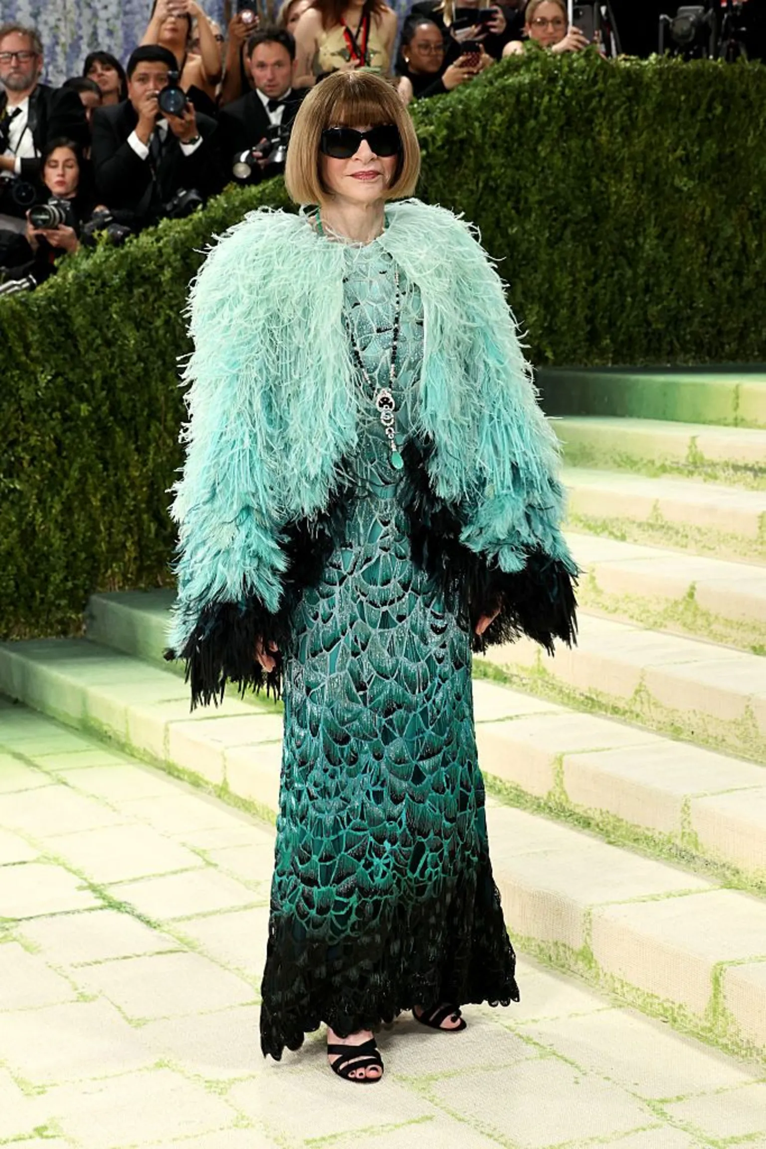  Anna Wintour wears her hair in her signature bob and sports dark sunglasses. Her dress is an avian-inspired confection, with a turquoise feathered cape on top of a darker-green dress whose embroidery resembles feathers.