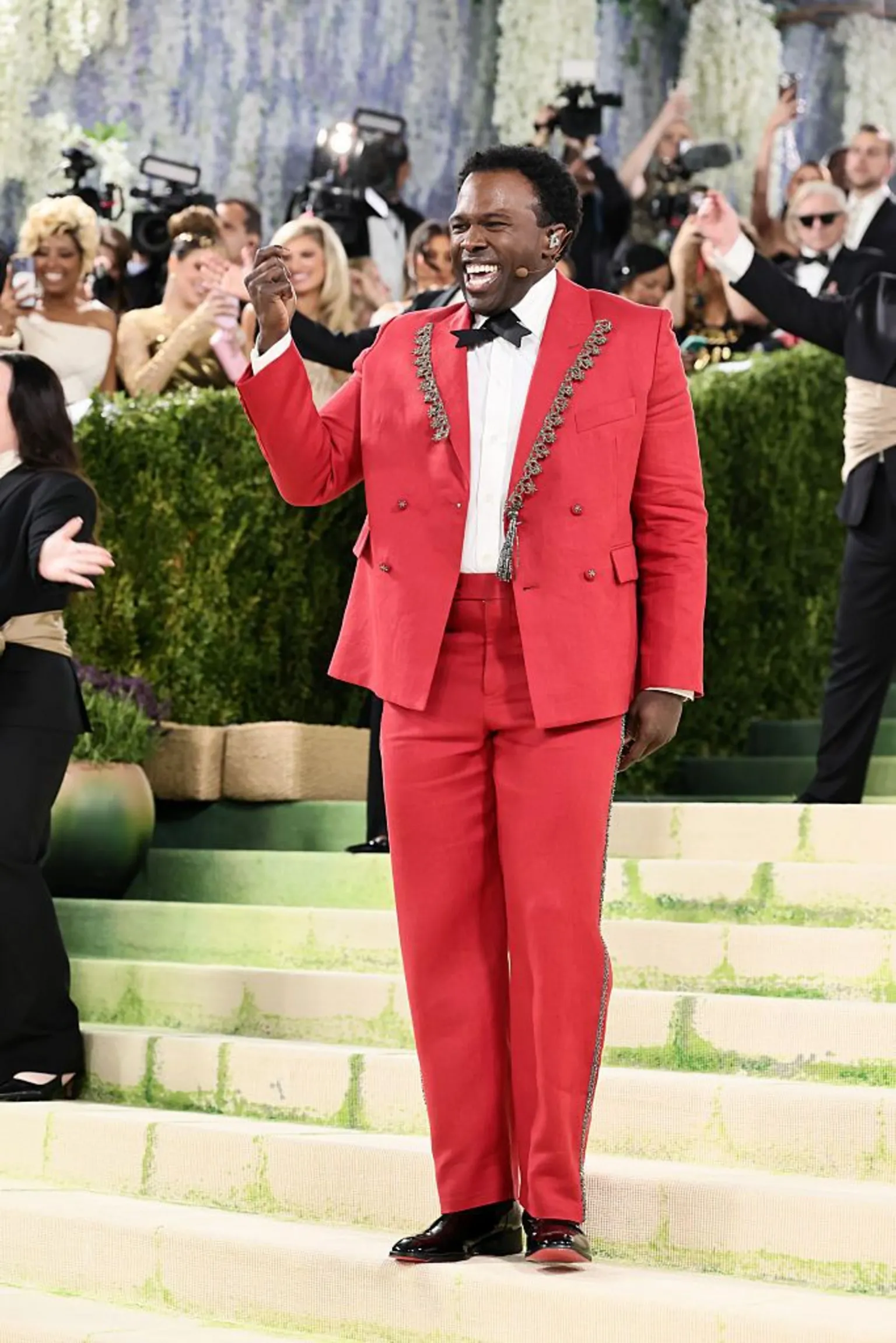  Broadway actor Joshua Henry wears a red double-breasted tuxedo with silver lapel embellishments. He stands on the stairs and sings as he performs for the press.