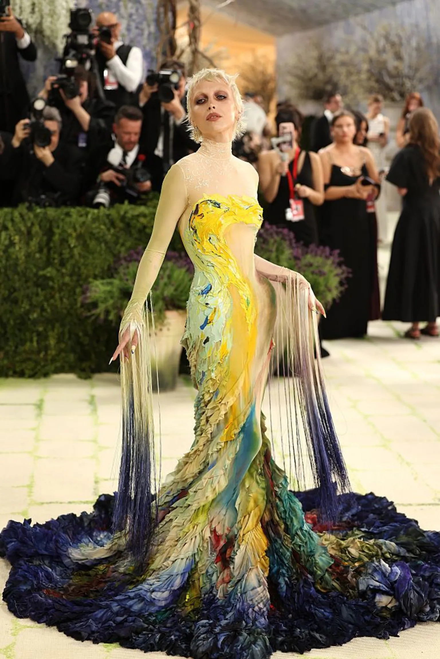  Influencer Emma Chamberlian wears a multi-colored gown that resembles a painting. It begins with dark blue colors at the base before transforming into a yellow and teal abstraction. Her sleeves also have fringes down to the floor.