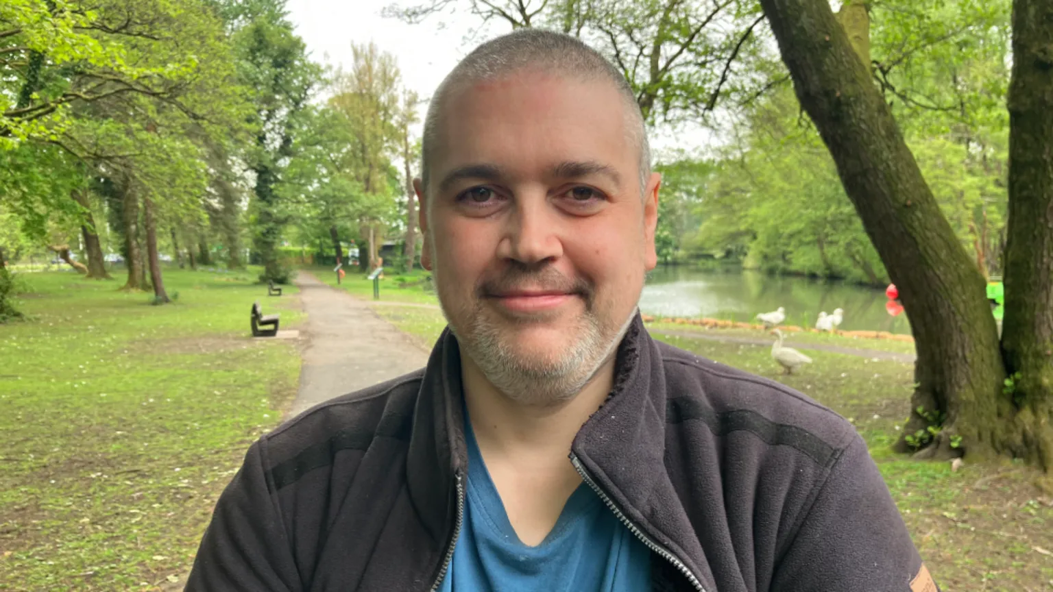 A man with extremely short grey hair and a grey beard and moustache. He is wearing a blue t shirt and black zip-up fleece and is stood in a green park with a river and trees behind him. He is looking at the camera and smiling.