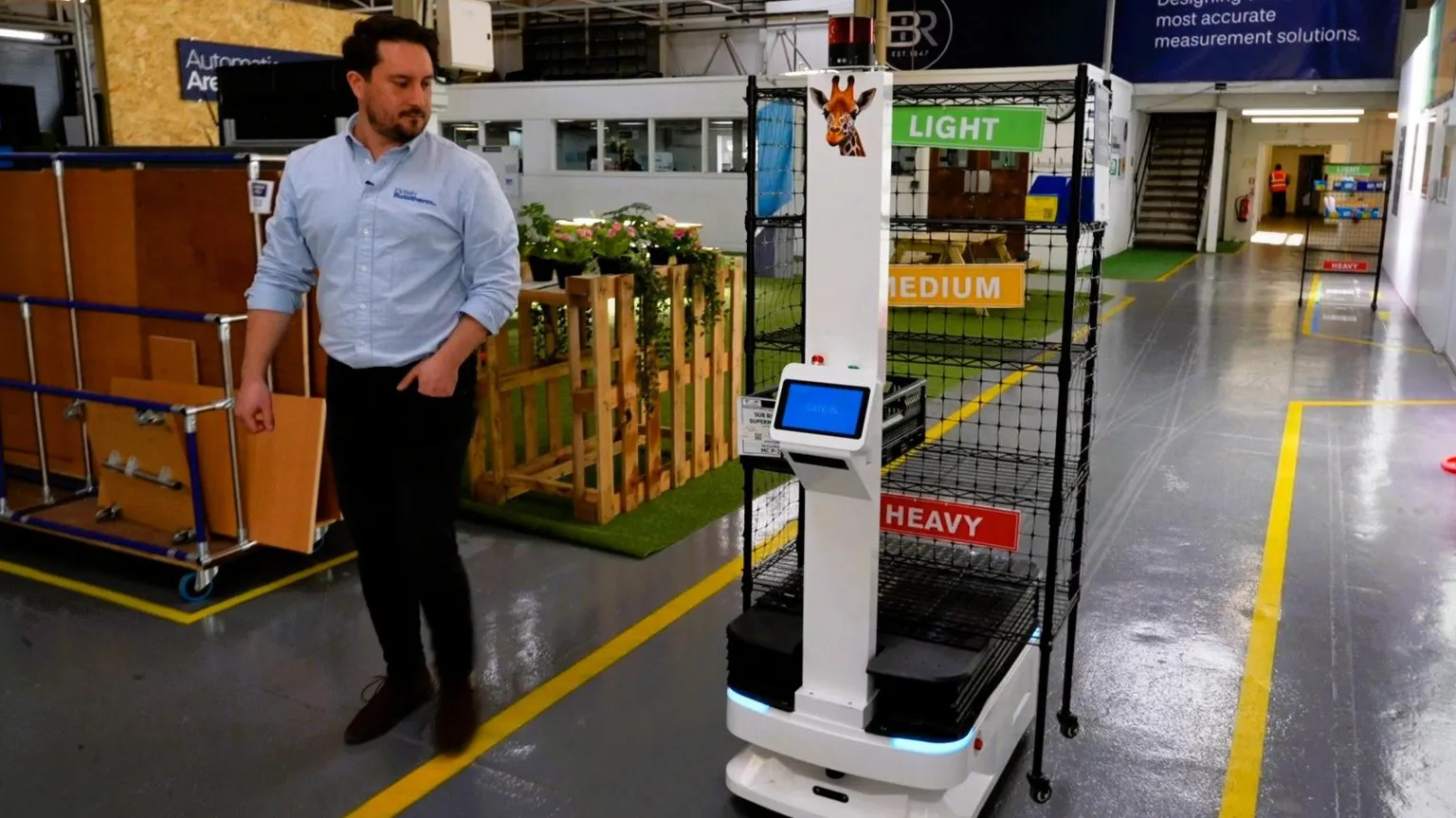 A robot, comprised of a floor-level platform and a tall white neck with a display screen, is seen in a factory environment. Yellow lines mark the path that the robot is following, while a worker stands to one side and is looking at the robot. The robot has a photograph of a giraffe attached to the top of the neck. It is carrying a black trolley with storage labels indicating where to store heavy, medium and light items.