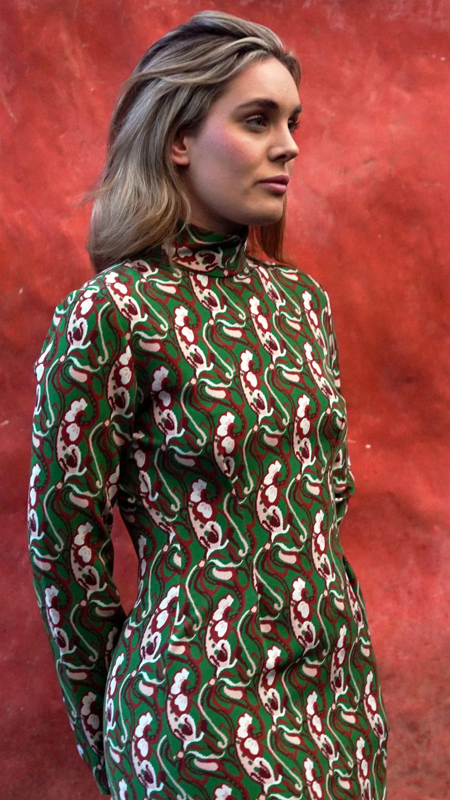 S4C Naomi standing in front of a large, textured red wall. She is wearing a green, red and white patterned slim-fit dress and is stood slightly to the side. The wall behind her fills the entire background and has variations in colour and surface texture.