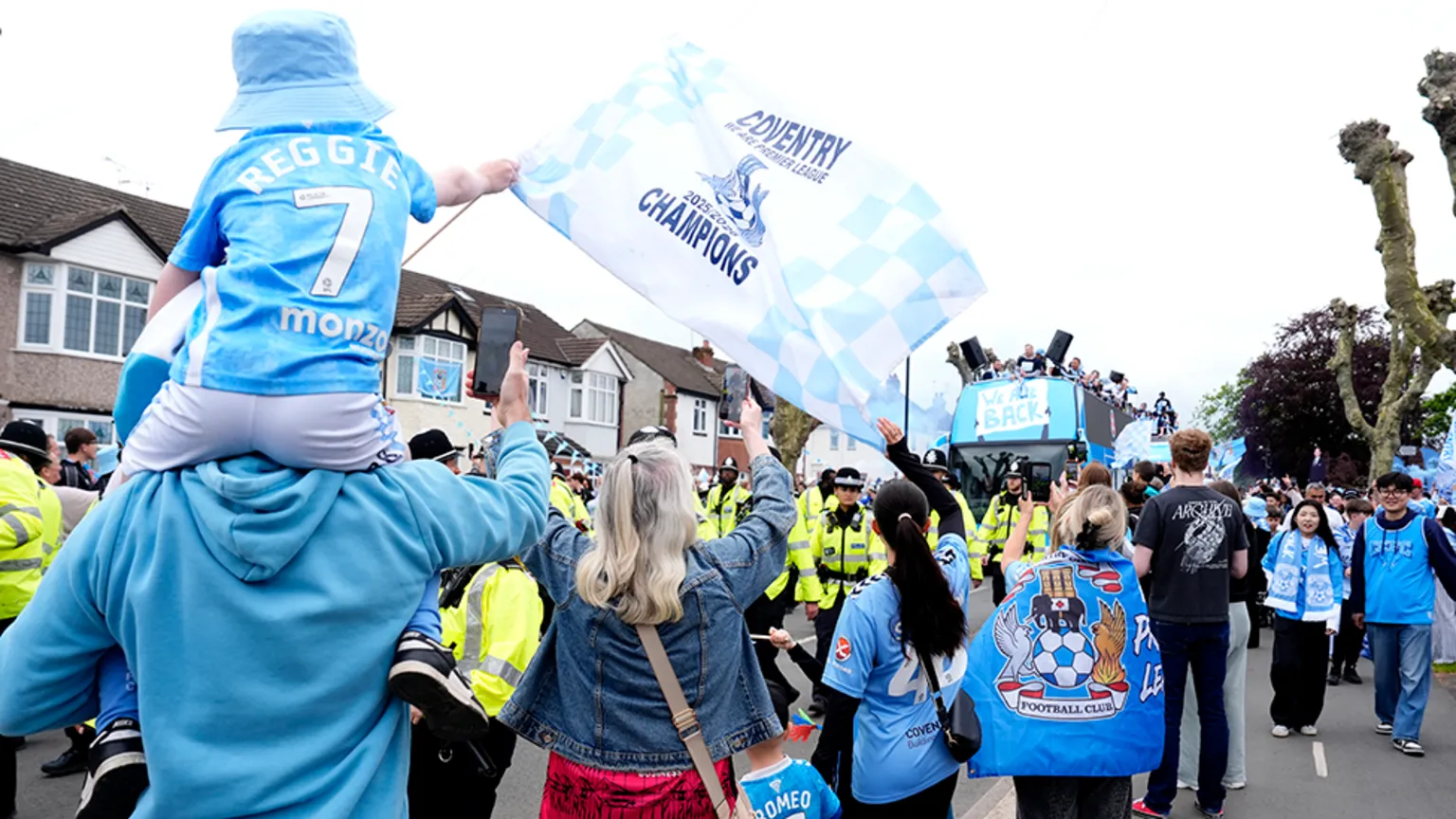  Fans lining the street as the bus approaches, with police in front of it in high-vis jackets. One young fan in the foreground on the left is on an adult's shoulders