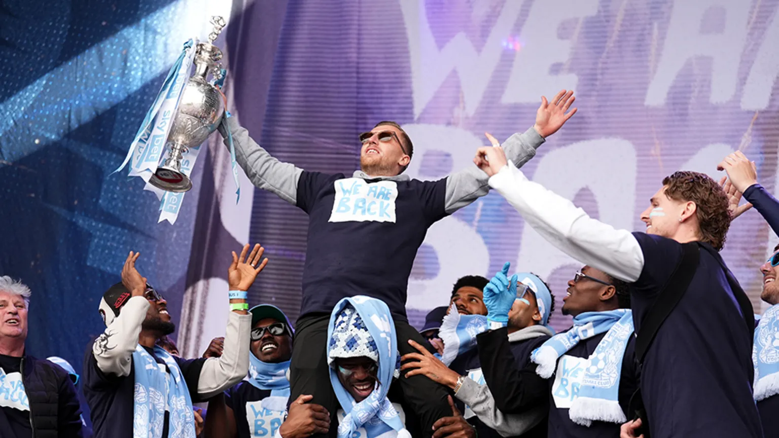  Jamie Allen is on a team-mate's shoulders, holding the Championship trophy aloft on stage. Lots of other players are in the background, smiling