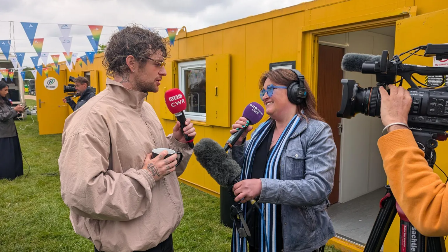 Tom Grennan is wearing a light brown jacket and holding a red BBC CWR microphone. He is speaking to radio presenter Lorna Bailey, who is in a denim jacket and holding two microphones. there is a TV camera on the right and yellow demountables and white marquees in background