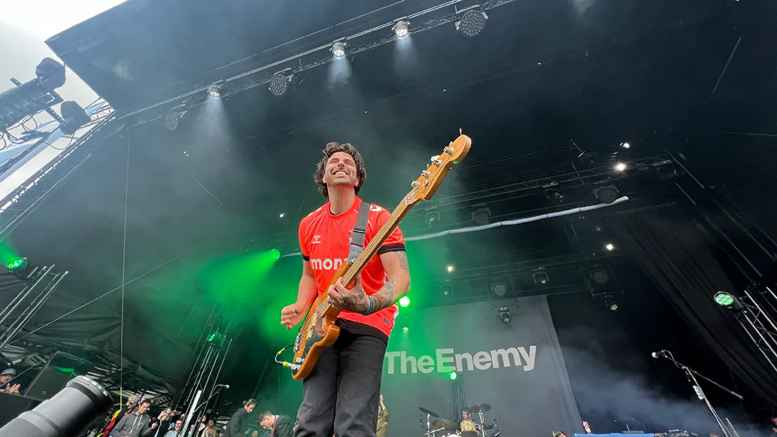 A member of the The Enemy holding a bass guitar and wearing a bright red t-shirt and blue jeans grinning on stage