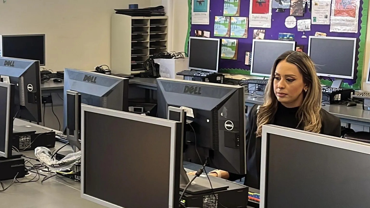 Woman sits behind a computer screen in a class full of screens - with a purple notice board behind her 