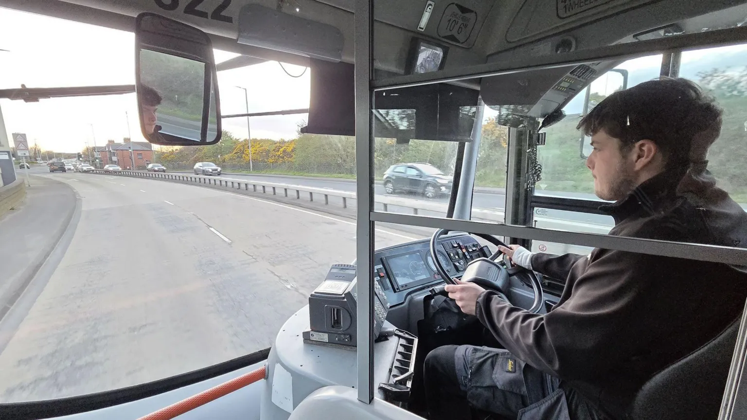 Luke Wilson Luke Wilson driving a bus. He has brown hair, wearing a black jacket and black trousers. He is driving along a dual carriageway with cars on the other side.