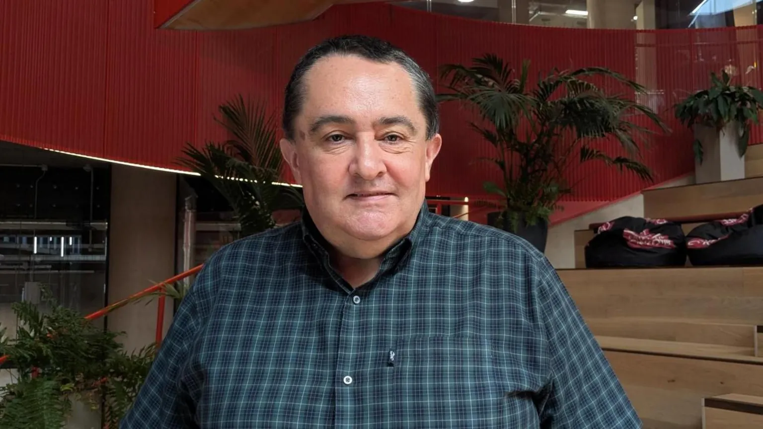 Martin's face is clearly visible and faces forward toward the camera. The hair is short, dark, and neatly combed, with a slightly receding hairline. The face is smooth and clean‑shaven. He is wearing a blue chequered shirt.