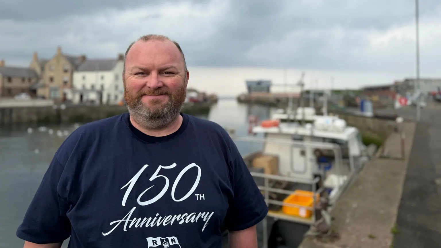 A short-haired balding man with a beard is standing in front of the harbour. if you look over his shoulder you can see right out to sea. He is wearing a navy t-shirt that reads '150th Anniversary'.