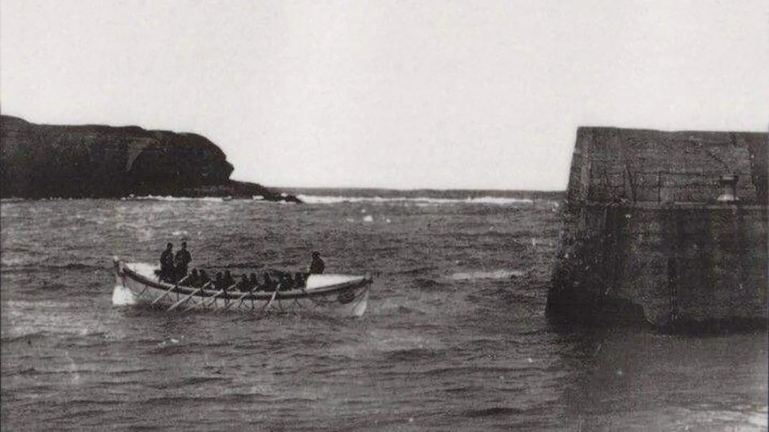 RNLI An old black and white picture showing a wooden longboat, there are 5 oars on the boat. they are just at the edge of the harbour.