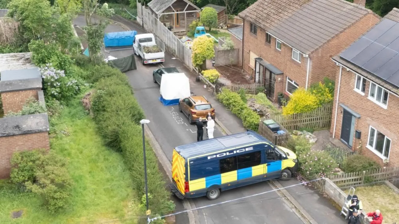  A drone shot above several homes and gardens - a residential road running through the middle which has a forensics tent, police van and sheets up to cover areas.