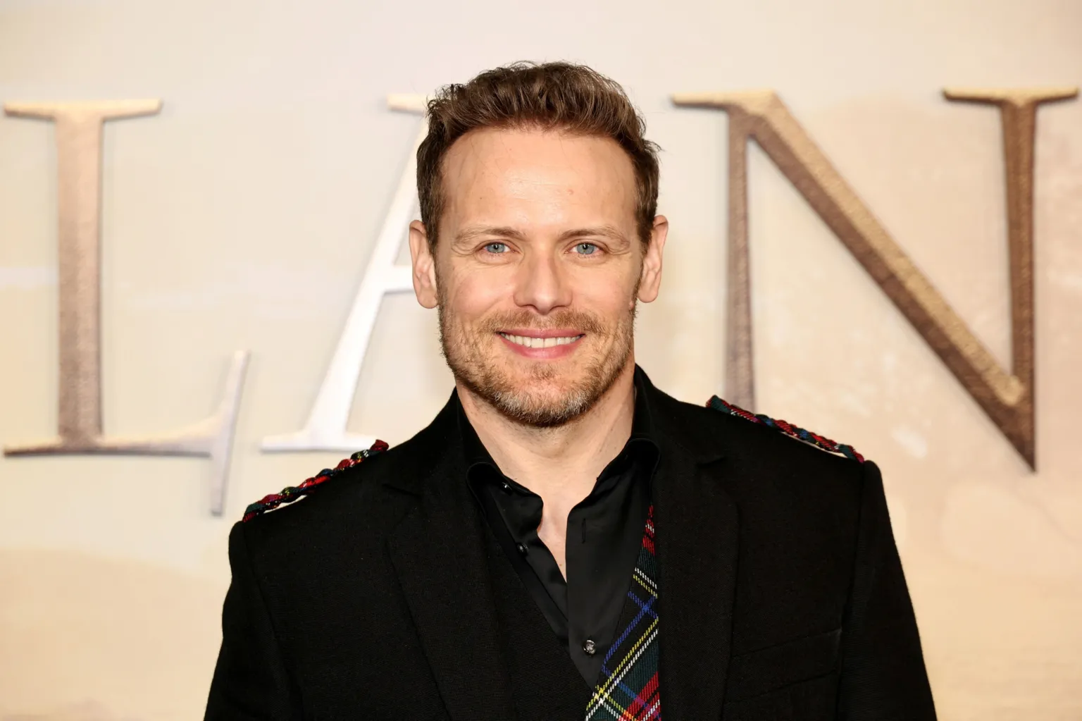  A man is smiling at the camera. He's wearing a black blazer and shirt it was tartan elements added to it