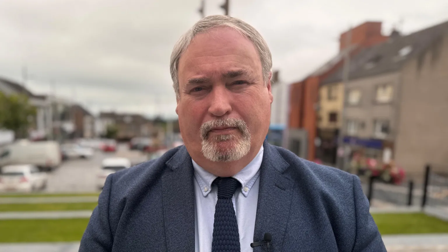 Colm Gildernew in a blazer and tie stands outdoors, with a blurred street and buildings behind him under a cloudy daytime sky.