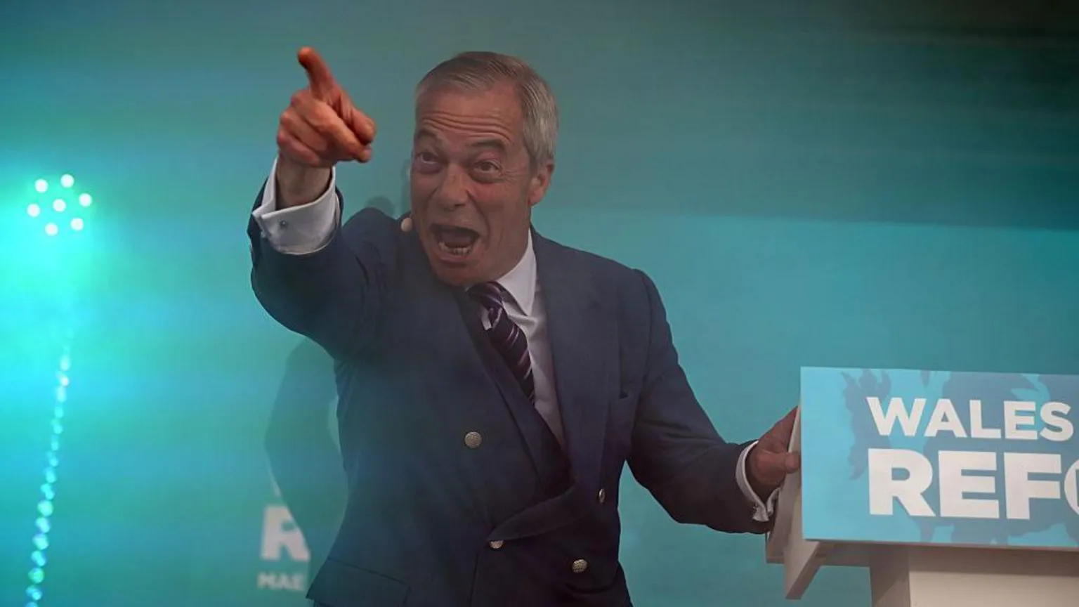  Nigel Farage stood at a lectern, pointing to the crowd and making a 