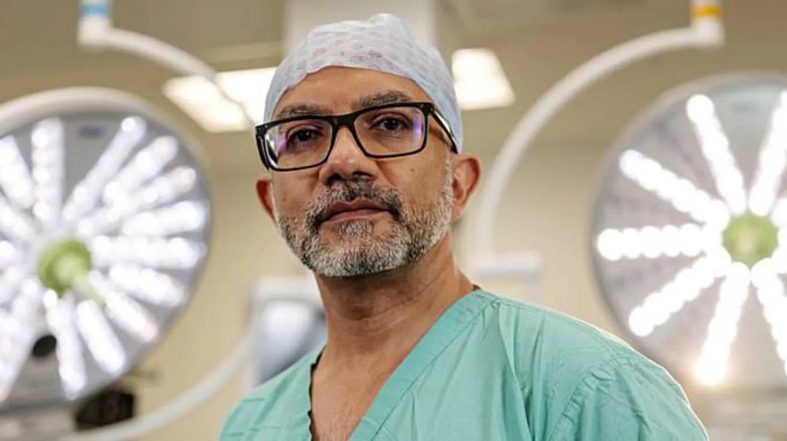 BBC/Dragonfly Film and TV Productions Surgeon CJ Shukla is standing with the large lights of an operating theatre behind him. He has a beard and is wearing surgical scrubs and dark glasses. He is looking into the camera. 