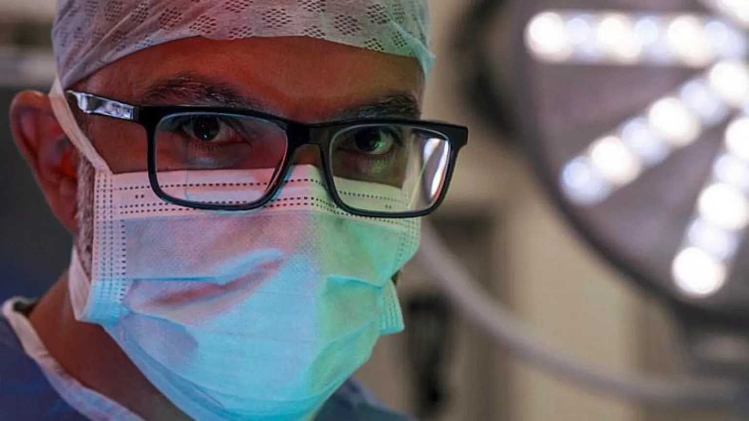 BBC/Dragonfly Film and TV Productions Close-up shot of surgeon CJ Shukla's face. He is wearing a surgical mask, scrubs and dark glasses and is staring into the camera. 