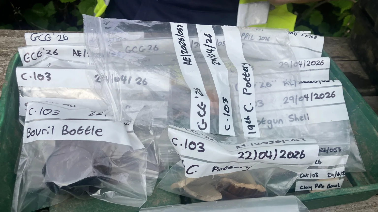 A green box holds roughly a dozen clear plastic bags. In each bag is a separate item with a label of what it is and a date. 
