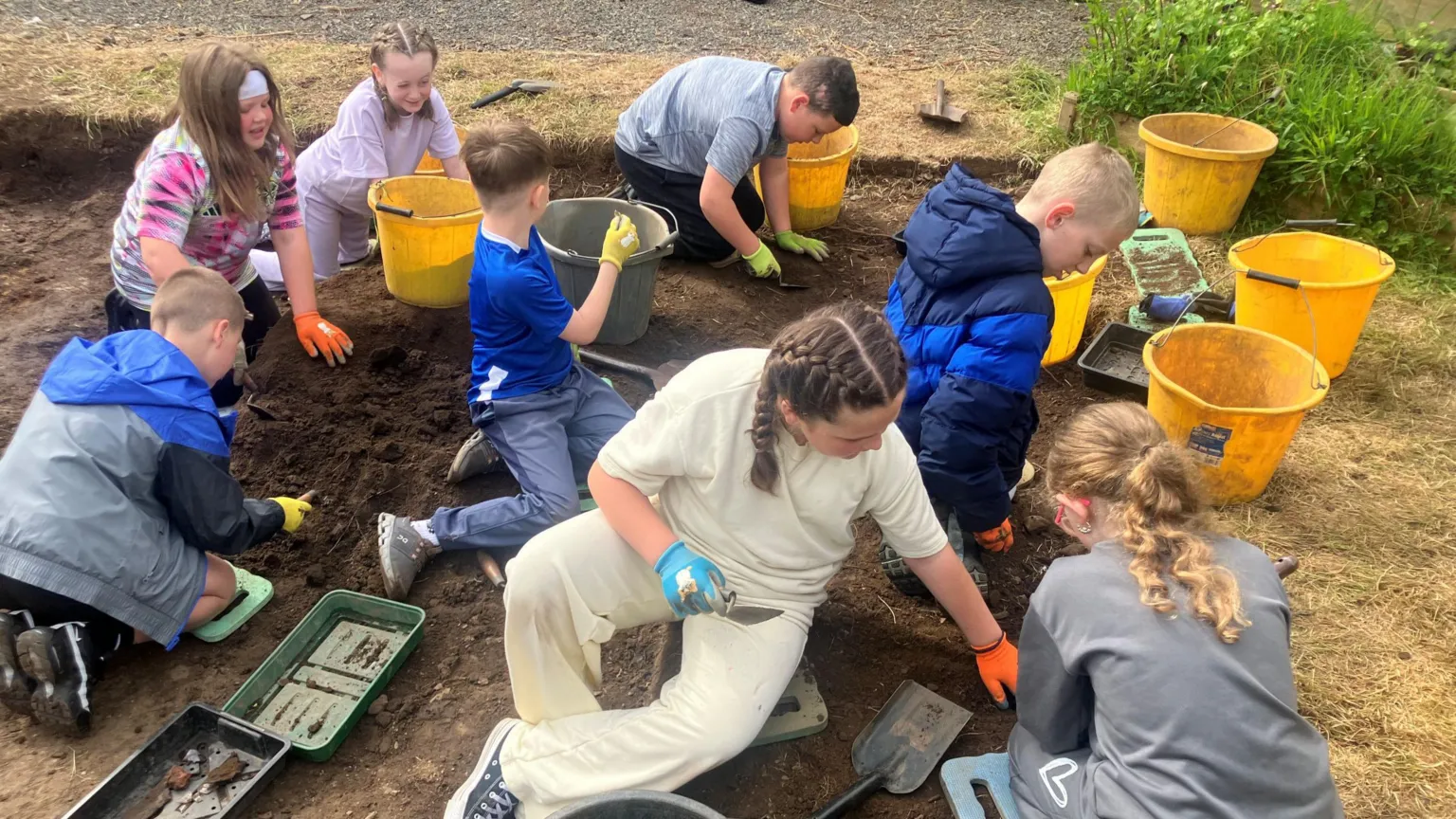 Eight school children kneel on the ground as they dig away at a patch of soil. They have each got trowels and a yellow bucket as they excavate the dirt. 