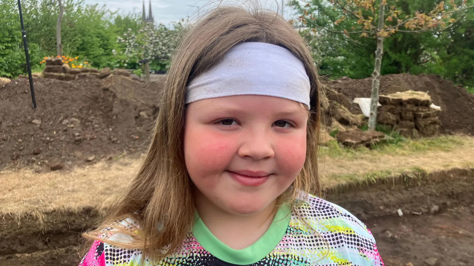 A girl with long brown hair wears a grey headband around her forehead. She wears a green, pink and white patterned T-shirt as she stares at the camera. 
