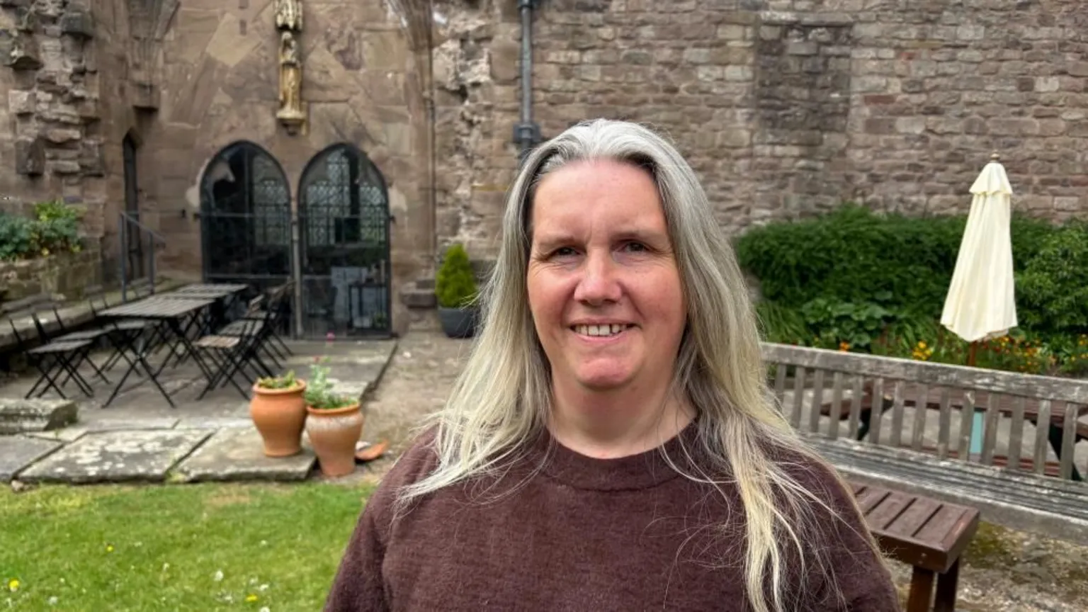 A woman in a cathedral garden with long grey hair wears a brown jumper. There is a set of tables and chairs and some plants in pots behind her.