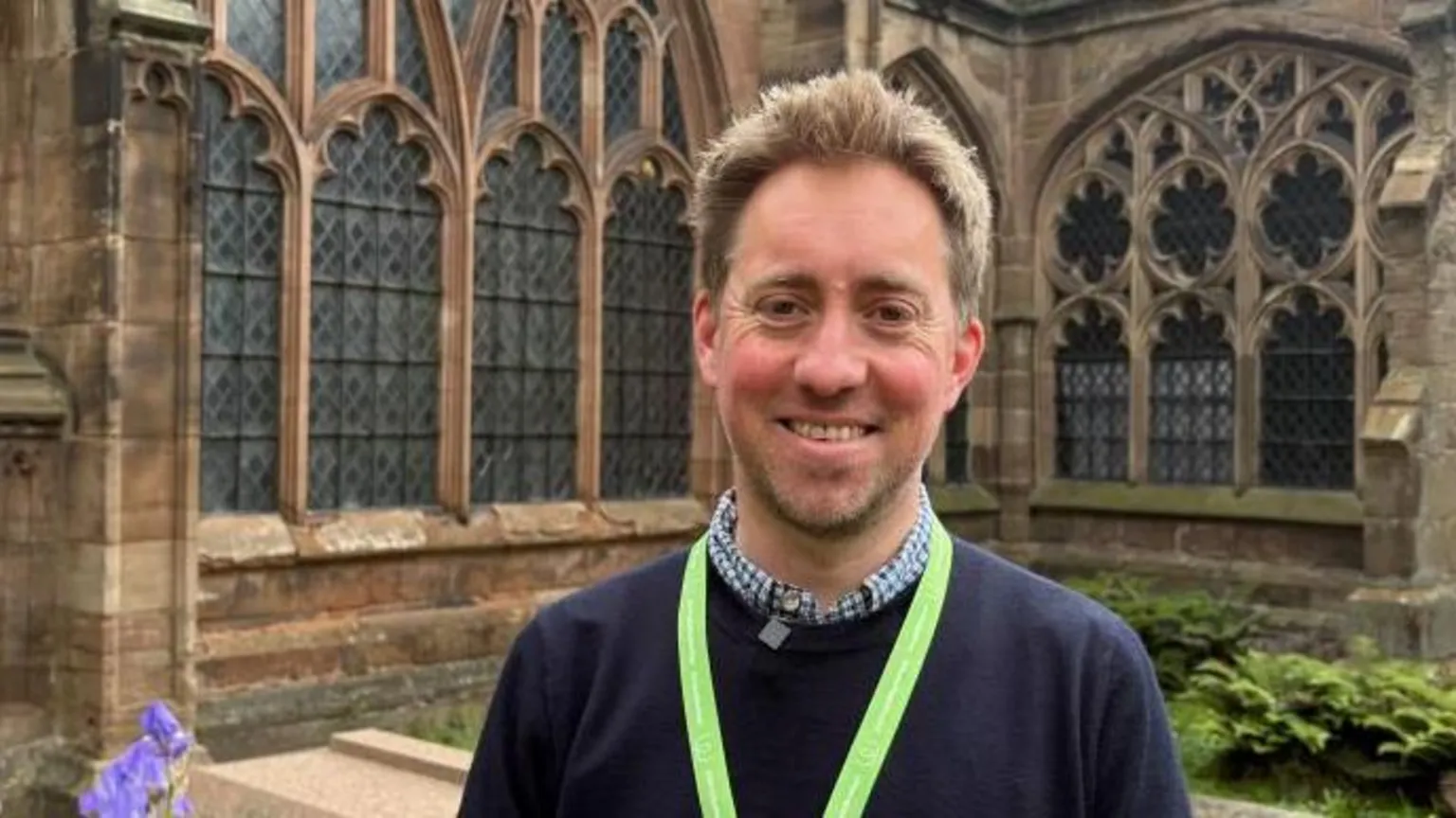 A man wears a checked shirt underneath a navy jumper with a yellow lanyard round his neck. He has short blond hair and is standing in front of a cathedral building with ornate leaded windows and stone walls.
