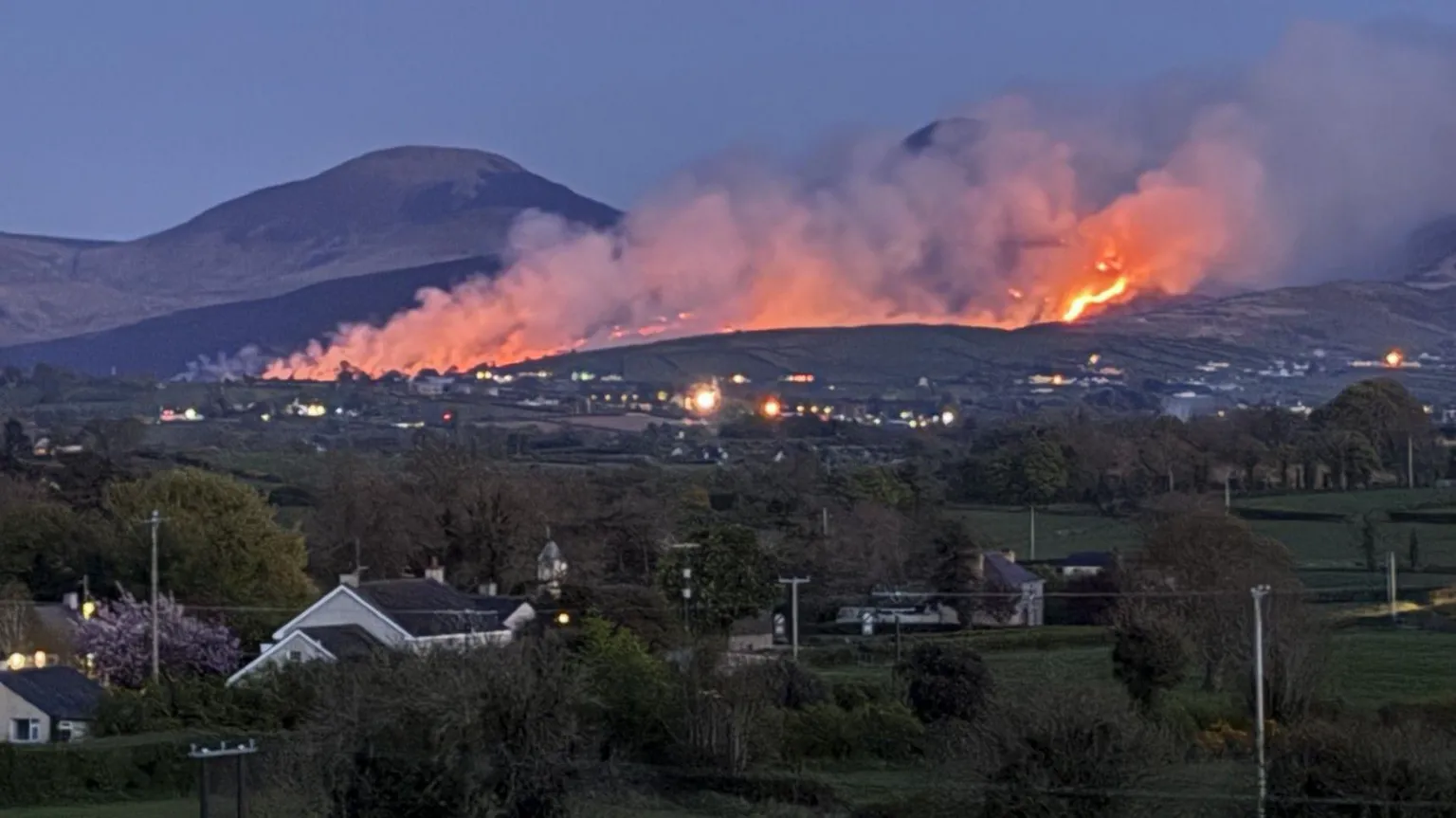 Beyond country houses, hedges, trees and rolling hills, a black mountaintop smoulders, with streaks of smoke and fire rising into the sky. 