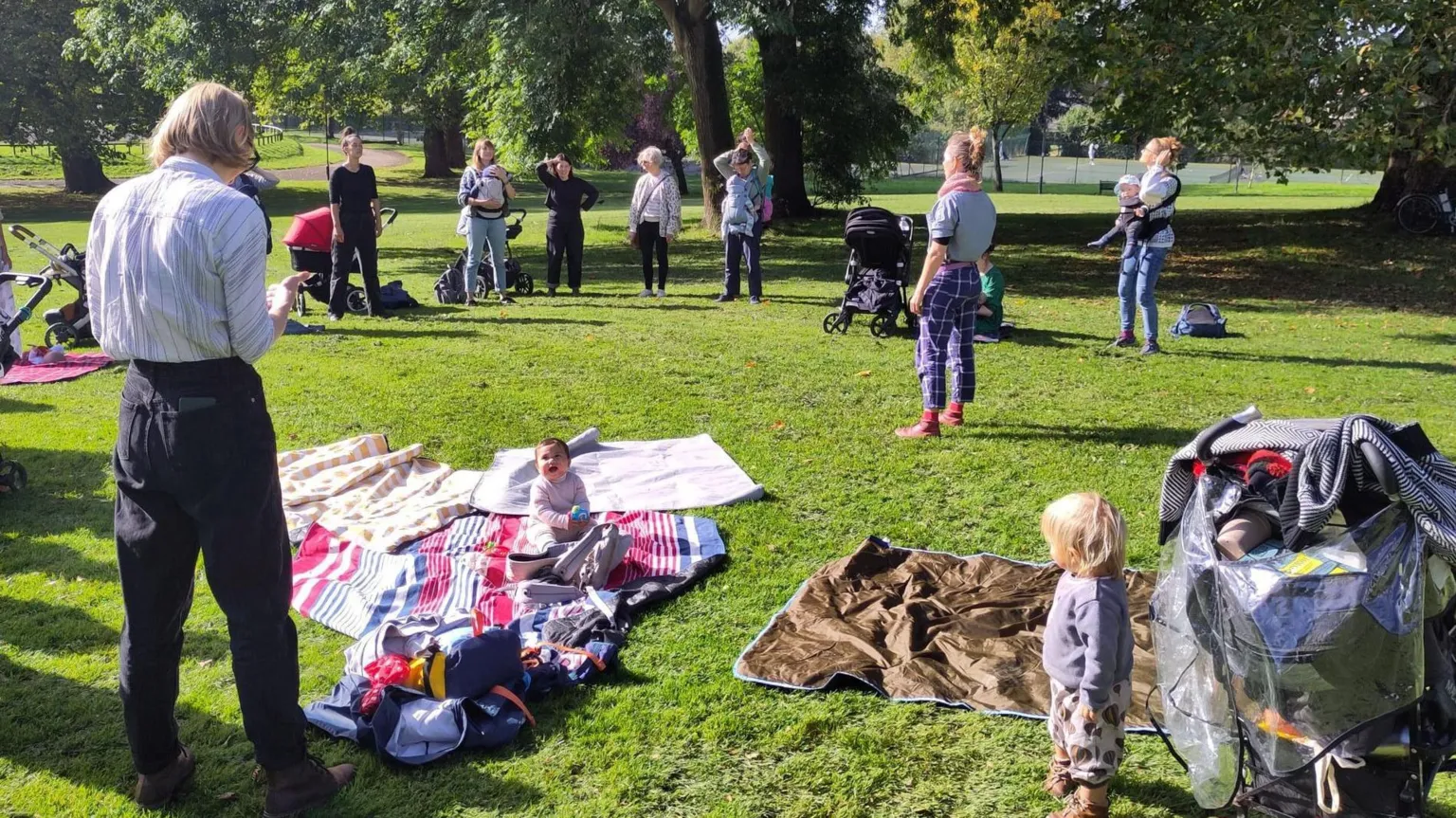 BYOB A group of people standing in a park on a sunny day. There are blankets on the floor and several children can be seen sitting or standing amongst the group. 