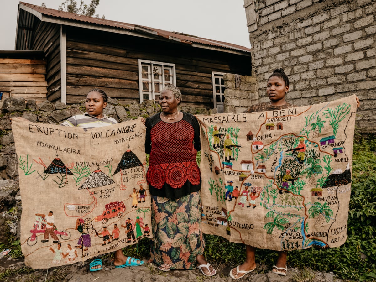 An older African woman stands outside a house as two younger women hold two large tapestries on either side of her