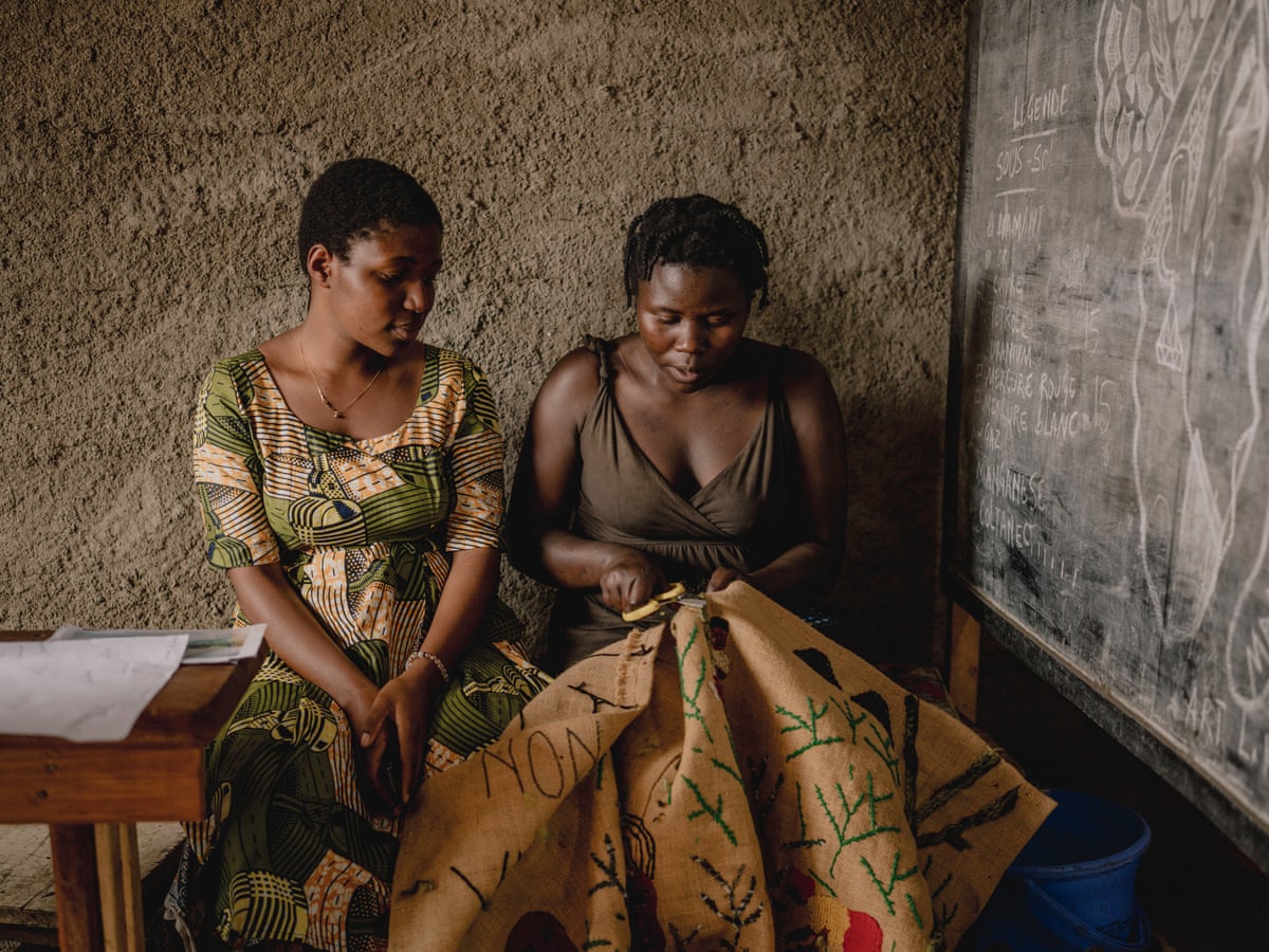 A young woman watches while another sews on a piece of sacking. 